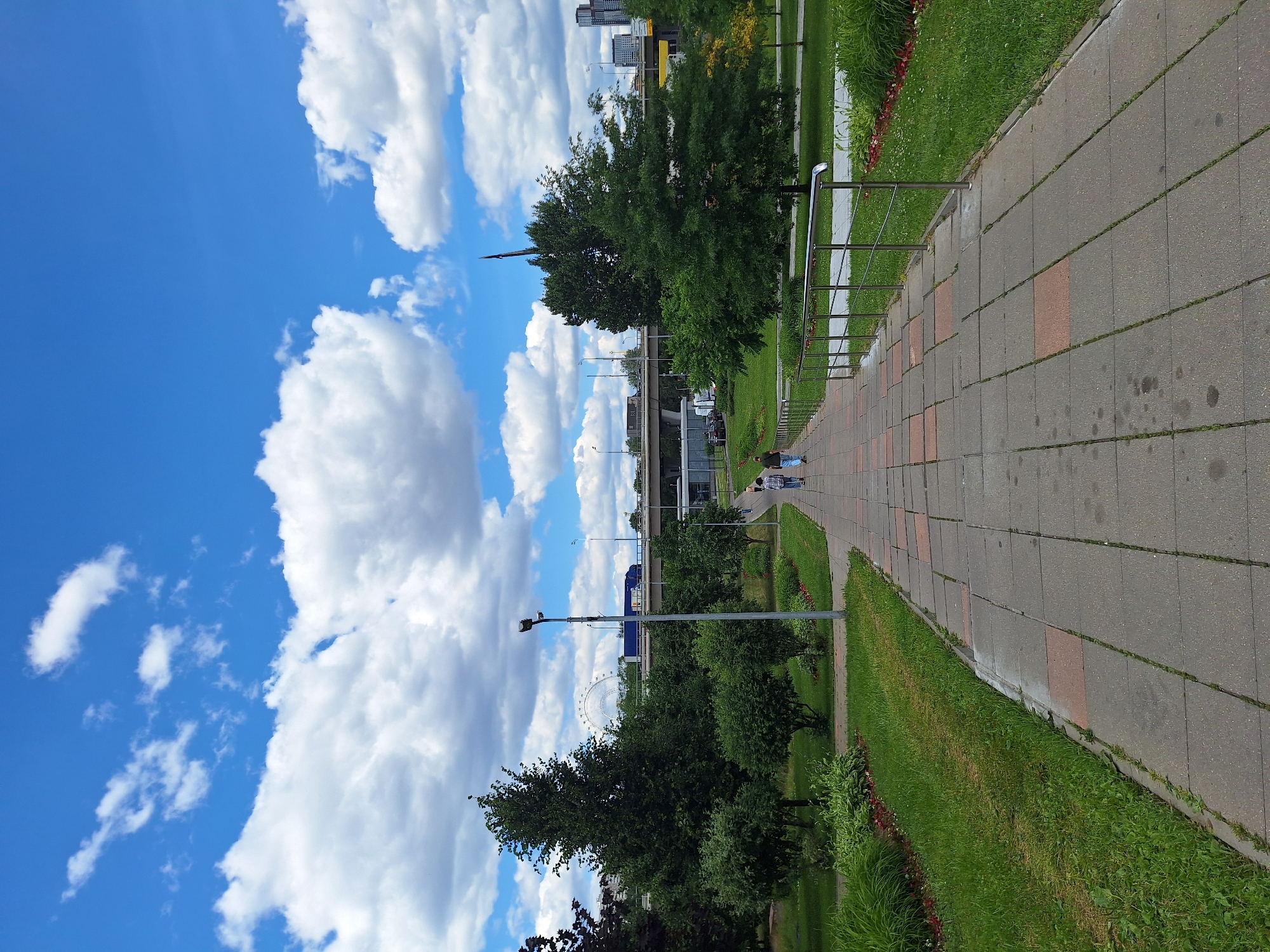 A vertical, low-angle shot captures a bright, sunny day in Moscow, Russia. The scene is a park with a paved walkway bordered by lush green grass and trees. The walkway is made of rectangular concrete slabs with smaller red square accents scattered throughout. A metal railing runs along the left side of the walkway, separating it from the grassy area.
In the distance, several prominent structures are visible: a Ferris wheel, a tall building, and a bridge. The sky is a vibrant blue, dotted with fluffy white clouds, indicating pleasant weather. Two people are walking on the paved path, their backs to the camera. One person is wearing a blue shirt and jeans, and the other is wearing a plaid shirt and dark pants.
The overall mood is serene and tranquil, suggesting a leisurely day in a city park. The presence of the Ferris wheel and the distinct architecture in the background hint at an urban park setting within Moscow. The lighting suggests it is daytime, with the sun casting light on the scene.