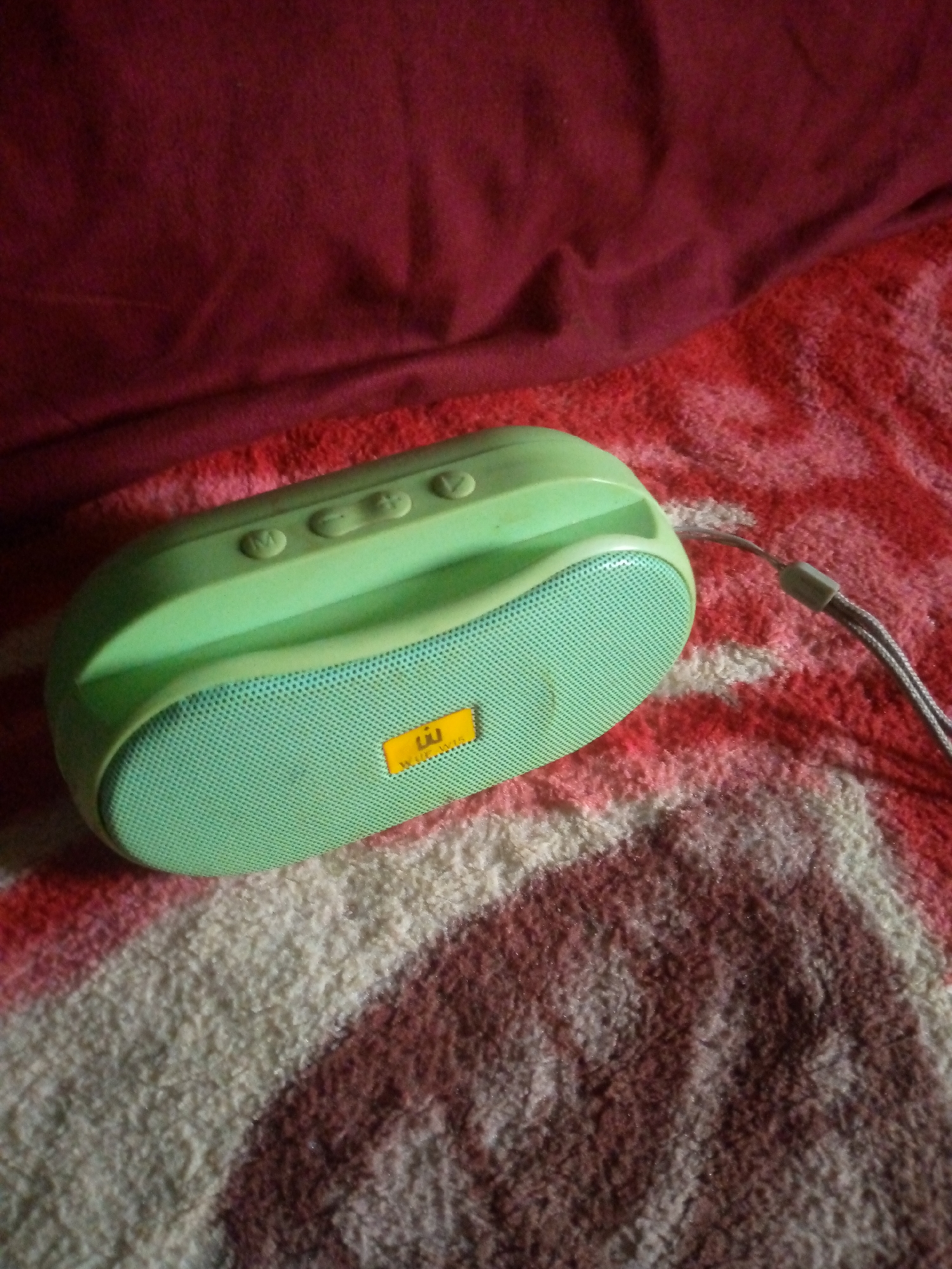 A light green Bluetooth speaker sits on a red and white patterned blanket, with a dark red fabric draped behind it. The speaker has a mesh front with a yellow sticker that has a black logo. There are several buttons on the top of the speaker, including what appears to be a power button and volume controls. A grey strap is attached to the speaker. The scene appears to be indoors, possibly on a bed. There are no people or animals visible in the image. The overall lighting is dim, suggesting it may be nighttime or the room is not well-lit. The image does not provide any specific context that would place it in Unguwan Rimi, Nigeria.