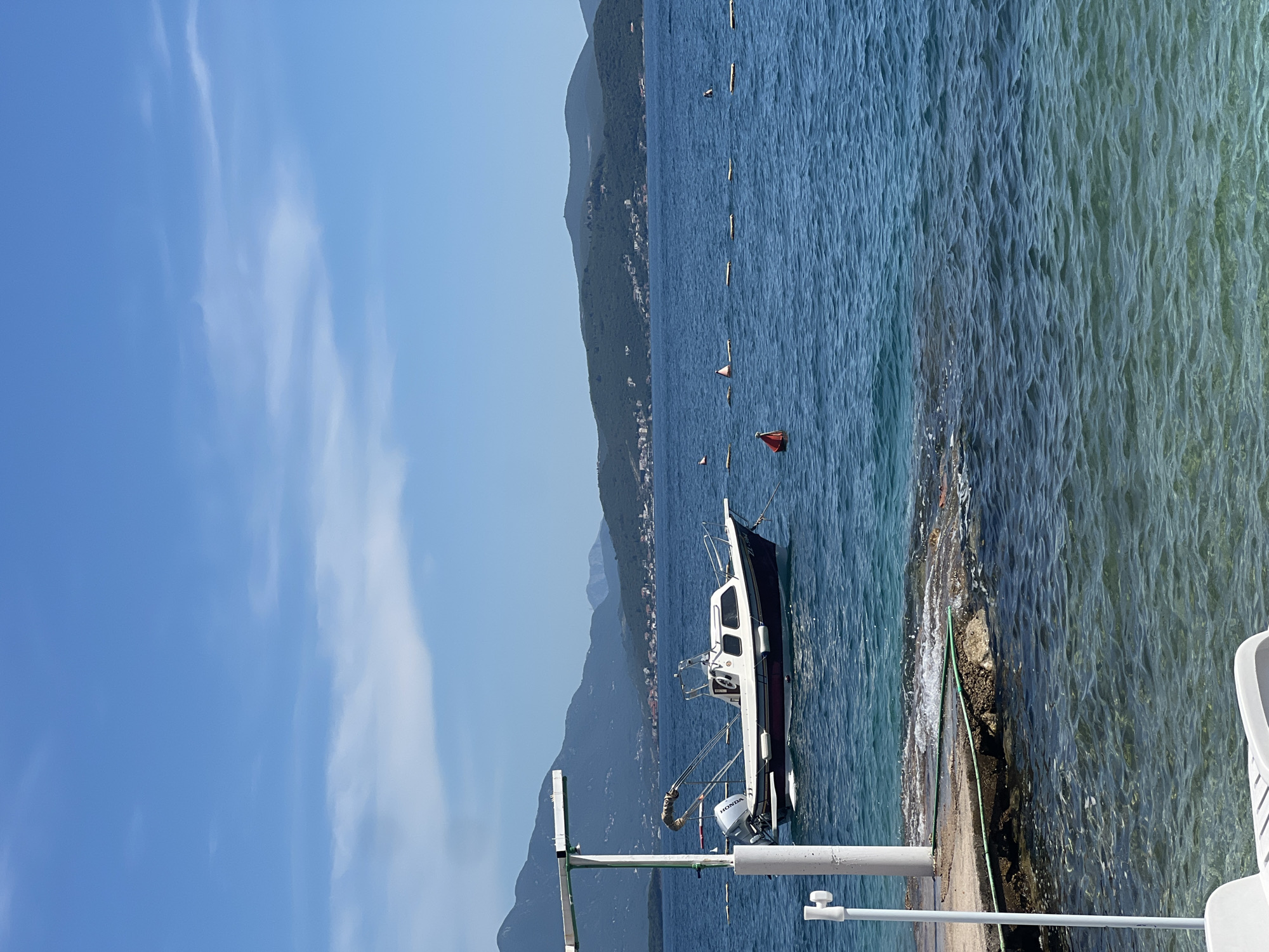 The image depicts a serene coastal scene in Baošići, Montenegro. A small motorboat, primarily white with a dark hull, is docked alongside a low concrete pier. The boat is equipped with fishing rods and what appears to be a small outboard motor. The surrounding water is clear and blue, with a line of buoys stretching across the bay, presumably marking a swimming area. In the background, a mountainous coastline is visible under a clear blue sky with scattered clouds. The overall impression is one of a peaceful summer day on the Adriatic coast.
