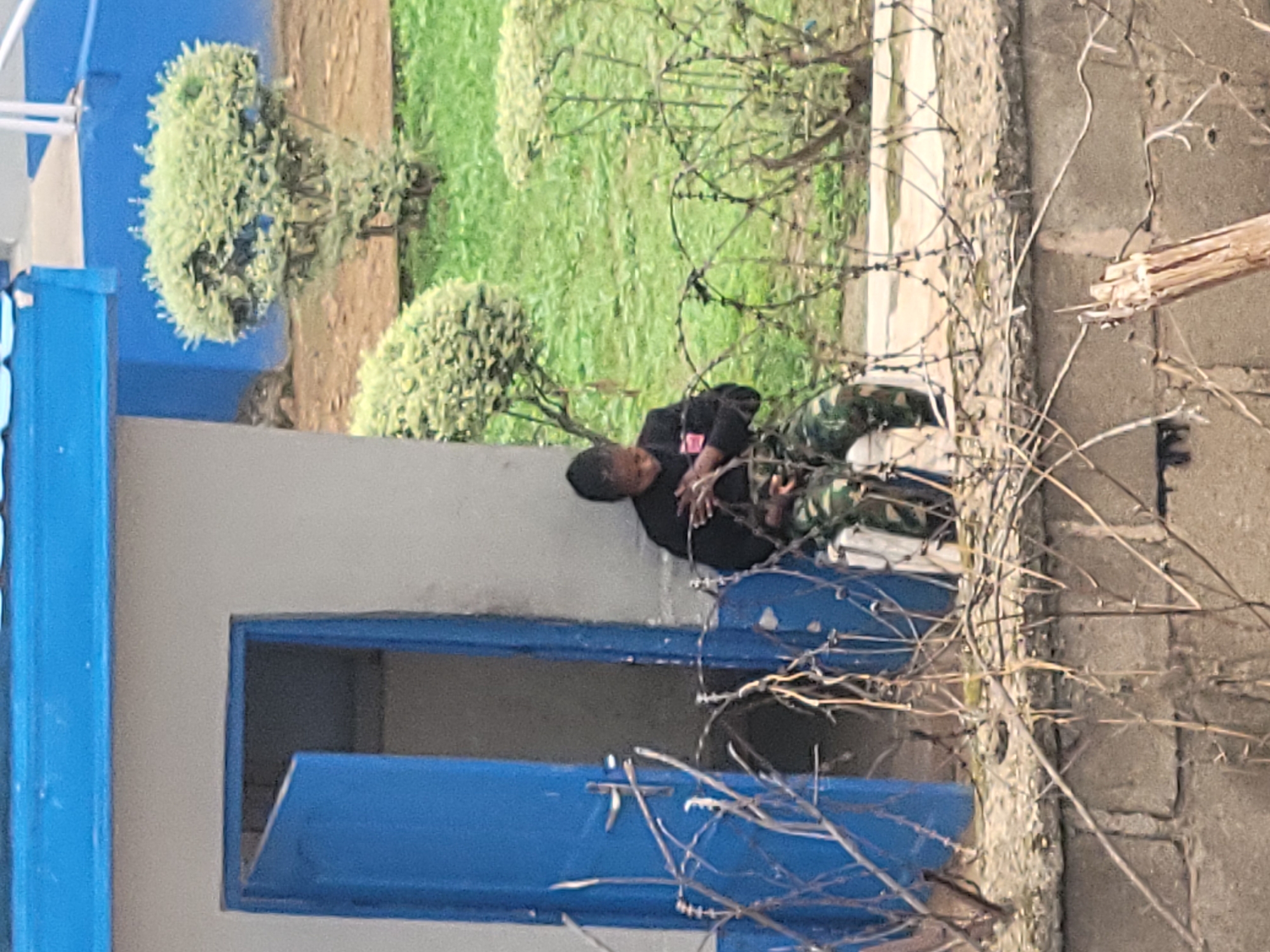 A person in camouflage pants and a dark jacket is sitting outside a blue-doored building in Gura Top, Nigeria. They are hunched over with their arms crossed over their chest. Barbed wire is in the foreground, and a well-maintained bush is behind the person. The building appears to be made of concrete and has a blue overhang. The ground is covered in grass and dirt.