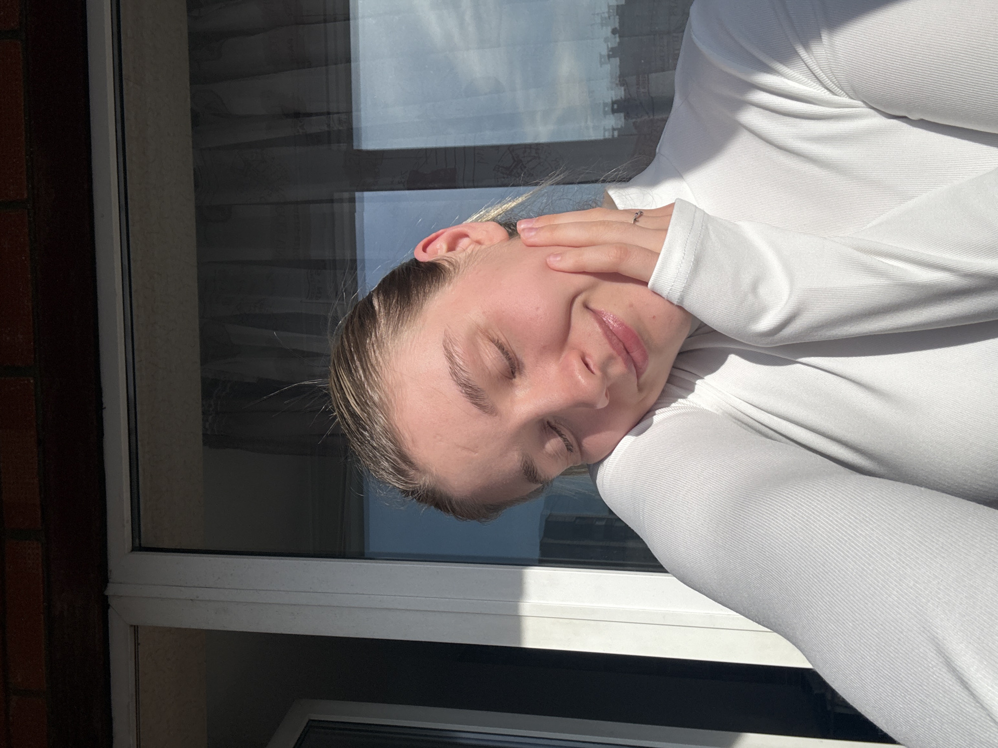 A young woman is positioned in front of a window. She is wearing a white, long-sleeved shirt. Her eyes are closed and she is smiling softly. Her right hand is placed on her cheek. The window is divided into multiple panes and reflects the sky and possibly buildings. The building is in Izhevsk, Russia. The location is unknown.