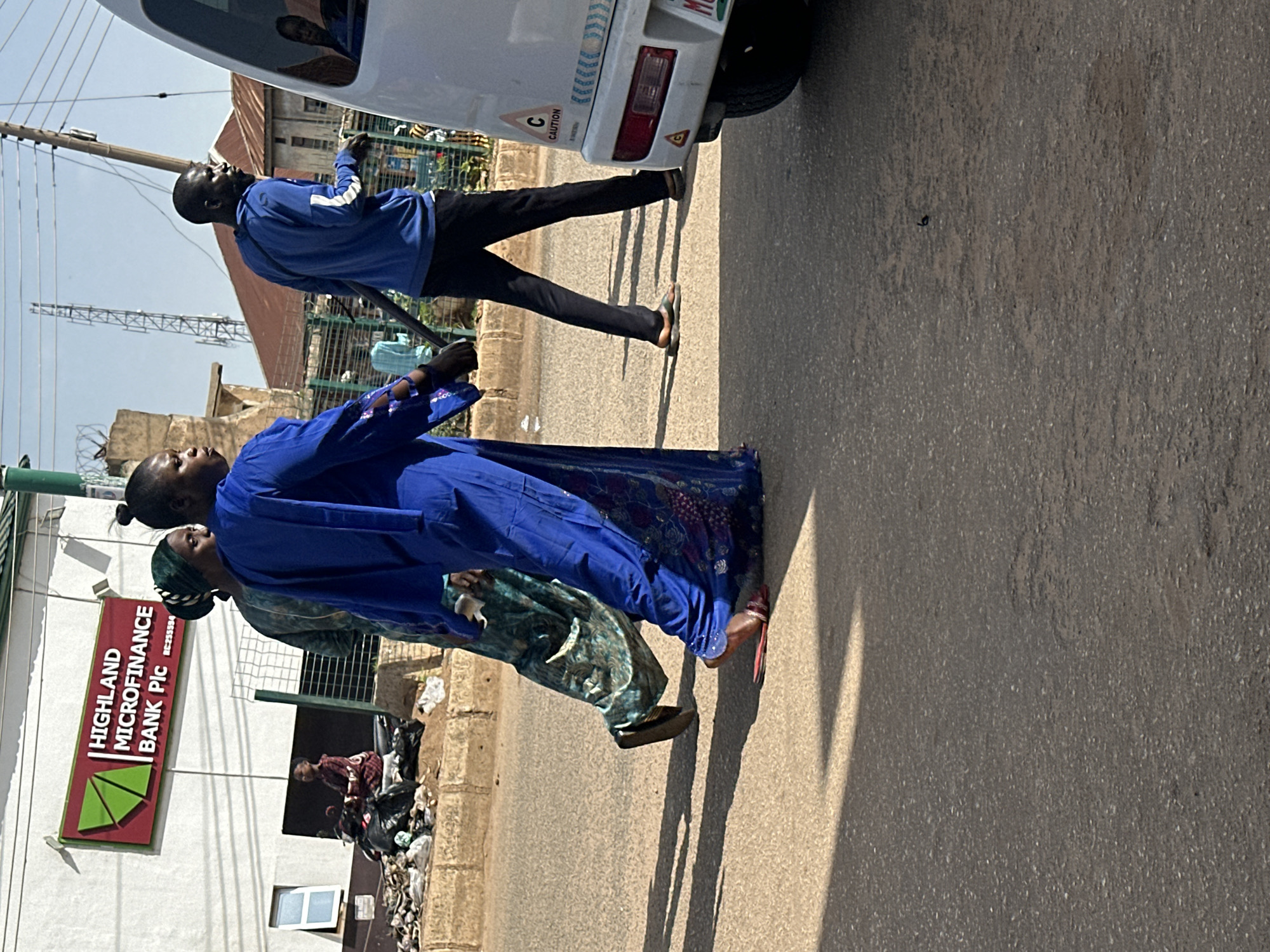 In Bukuru, Nigeria, two women wearing traditional blue and green garments walk on a paved road. A man in a blue jacket and black pants walks beside them, carrying a dark pole. In the background, a white van with caution signs and a license plate is parked on the side of the road. A building with a red and green sign that reads "HIGHLAND MICROFINANCE BANK PIC" is visible behind the people. The scene is illuminated by sunlight, casting shadows on the road.
