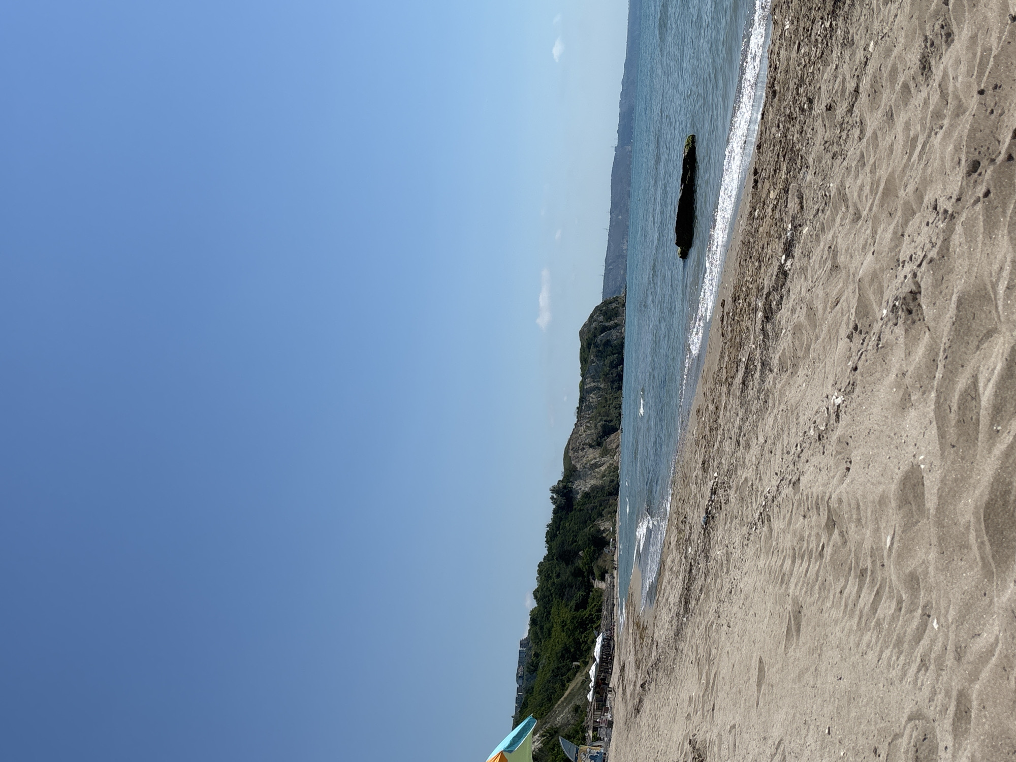 This is a landscape photograph of a beach in Topola, Bulgaria, under a clear blue sky. The foreground is dominated by a sandy beach with tire tracks impressed into the sand. To the left, a colorful beach umbrella with orange, green, and blue sections is visible. Further along the beach, a row of white umbrellas and what appear to be beach chairs or loungers are set up, suggesting a resort area. Dense green vegetation covers the slopes of a rocky cliff that rises from the sea on the left side of the image. The sea is calm with gentle waves breaking on the shore. A dark, moss-covered rock protrudes from the water in the middle ground. In the distance, the coastline stretches out, with hills and structures visible on the horizon. A few people are scattered along the beach and in the water.