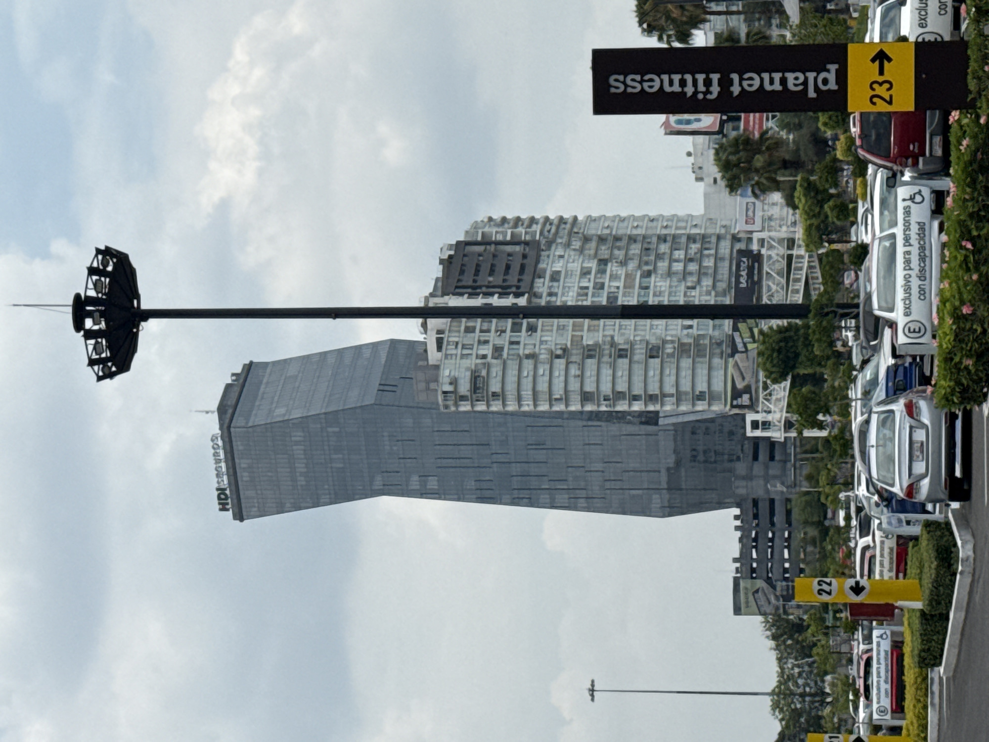 The image shows a parking lot next to a street in Puebla, Mexico with multiple skyscrapers in the background. Parking spaces reserved for people with disabilities are visible. The street is marked with parking lot direction signs indicating the aisles with numbers 22 and 23. A Planet Fitness sign is also present, along with "HDI seguros" written on the side of a skyscraper. A tall light pole stands to the left of the image. The sky is overcast.