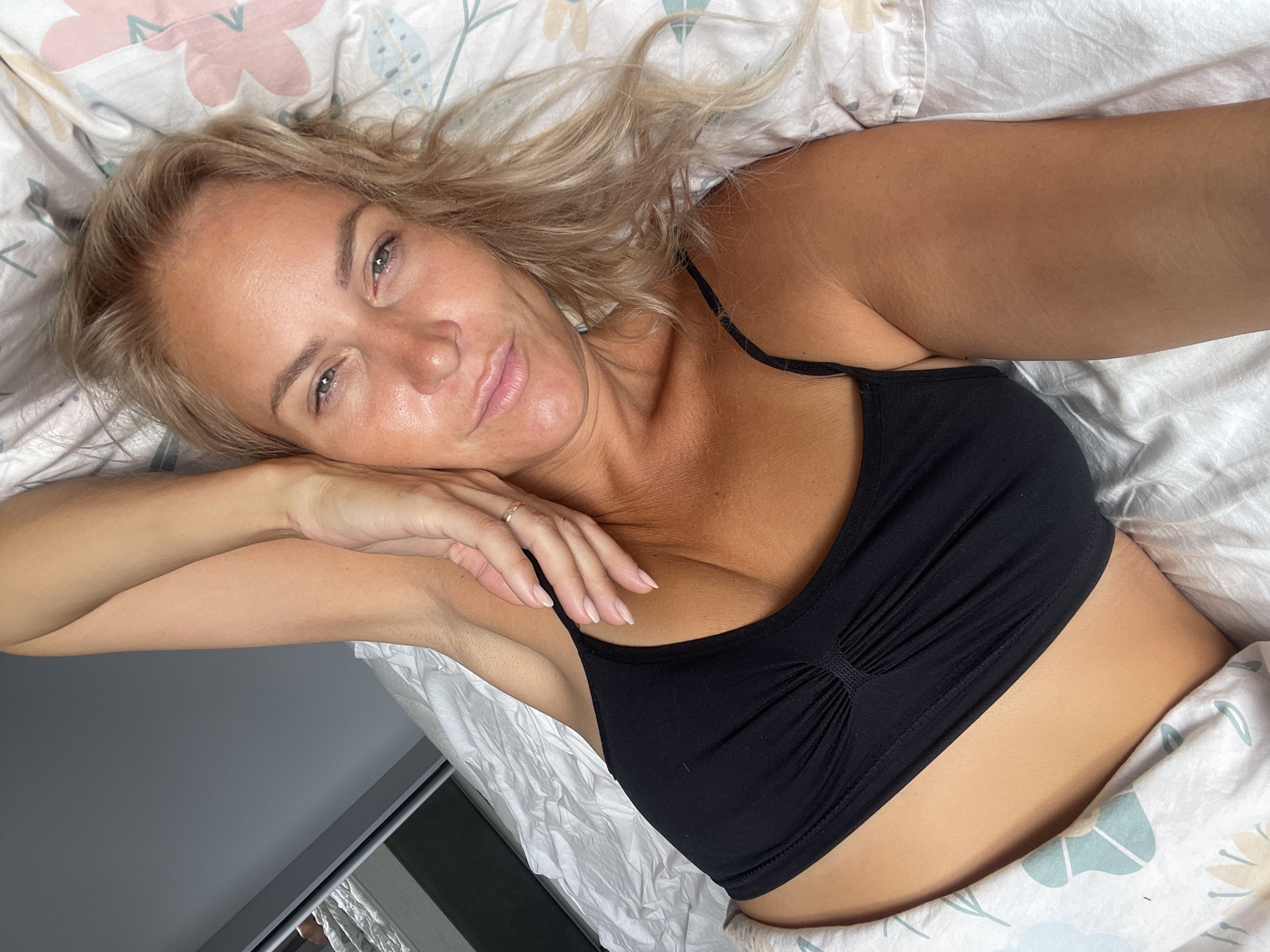 A blonde woman with fair skin and light blue eyes is lying on a bed. She is wearing a black tank top. Her right arm is bent at the elbow and rests on her head, with her hand touching her face. A ring is visible on her right ring finger. Her left arm is under the covers. The bed is covered with white and floral-patterned bedding. The lighting suggests it is daytime. The scene is indoors. There is no indication of Baranovka, Russia.