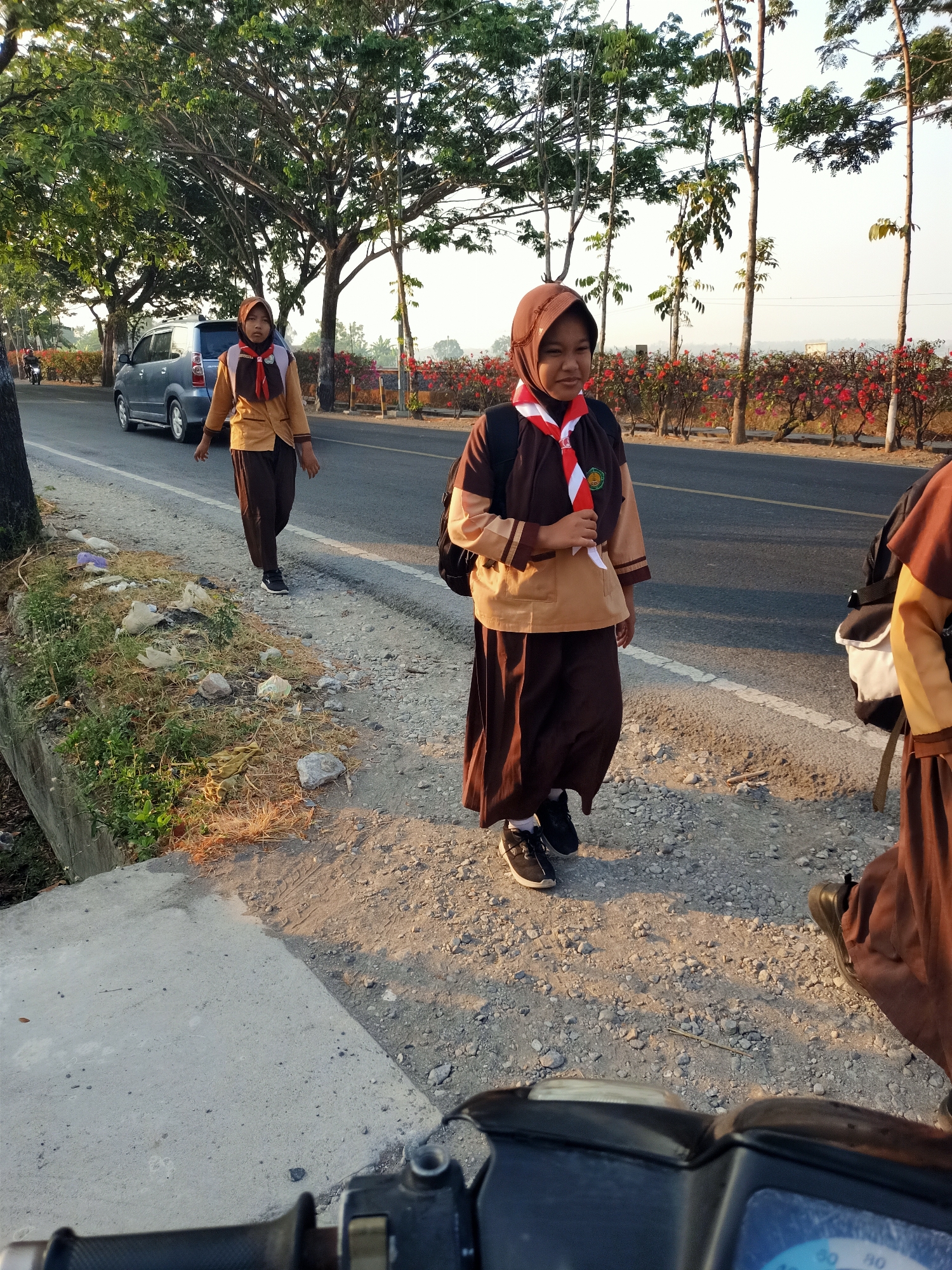 In Kendung, Indonesia, two young Indonesian female students in brown and tan uniforms with red and white neck scarves walk along a paved road during daylight. The student in the foreground, facing the camera and slightly to the right, is looking to her left. She has a dark backpack on. To her left, another student in a similar uniform walks away from the camera. She is also carrying a dark backpack. A silver minivan is parked on the side of the road to the left of the students. Trees with green leaves line the side of the road, and a line of flowering bushes with red blossoms runs along the far side of the road. The ground beside the road is a mixture of dirt, gravel, and sparse vegetation. The overall scene suggests a school day commute.