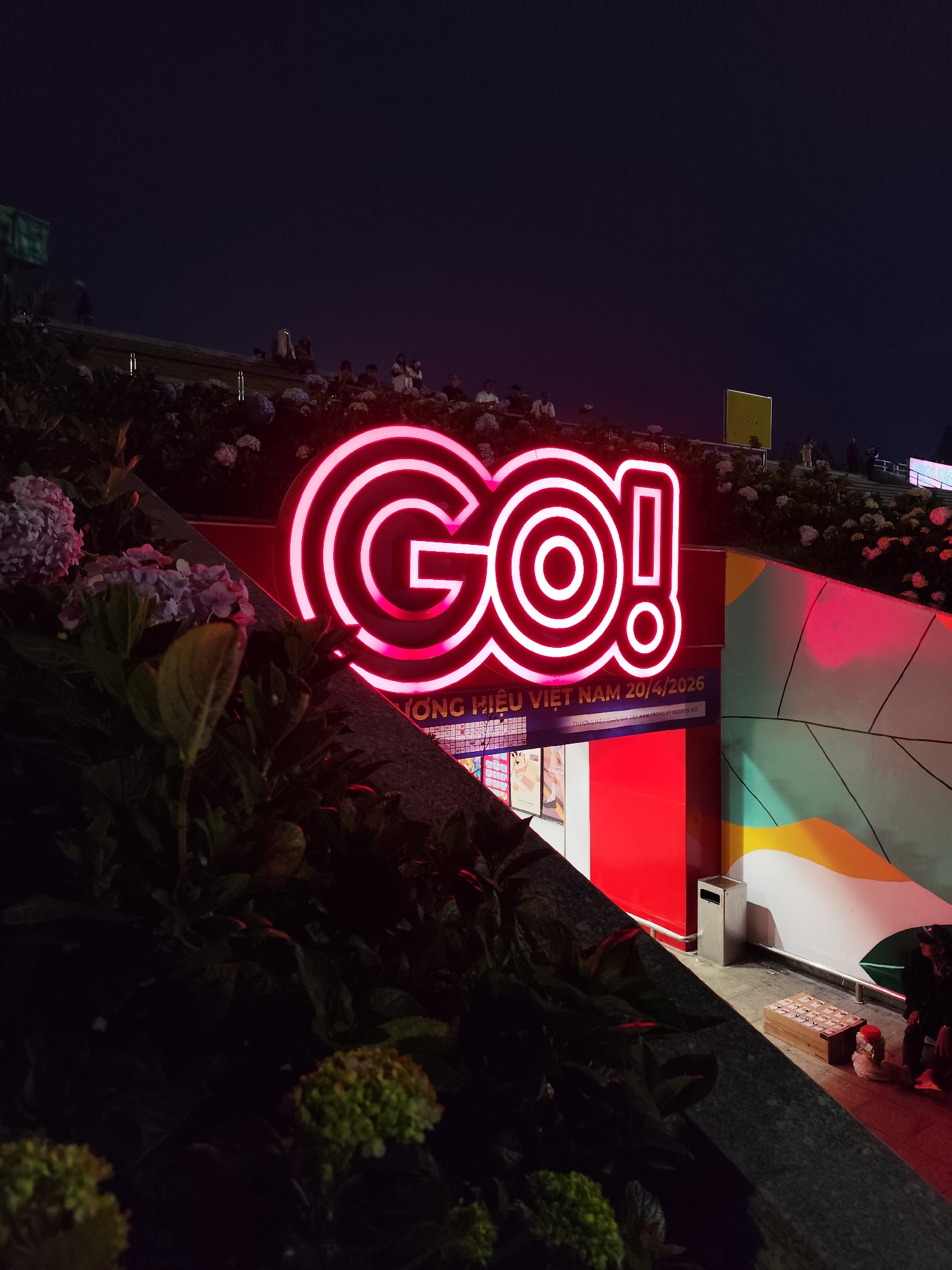 The image captures the exterior of a "GO!" store at night, prominently displaying a vibrant pink neon sign with the store's name. The signage includes Vietnamese text, including the date "20/4/2026" and the phrase "THƯƠNG HIỆU VIỆT NAM," suggesting it may be a promotional campaign or event linked to a Vietnamese national brand. The store has a modern design, with red paneling and a colorful mural adjacent to the entrance. A trash receptacle is visible near the store's entrance. In the foreground, a stone wall is covered with various flowers. In the background, people are gathered above the store on what appears to be a terrace. The image suggests a bustling evening scene, possibly taken in the city of Da Lat, Vietnam, known for its flower gardens and vibrant night atmosphere.