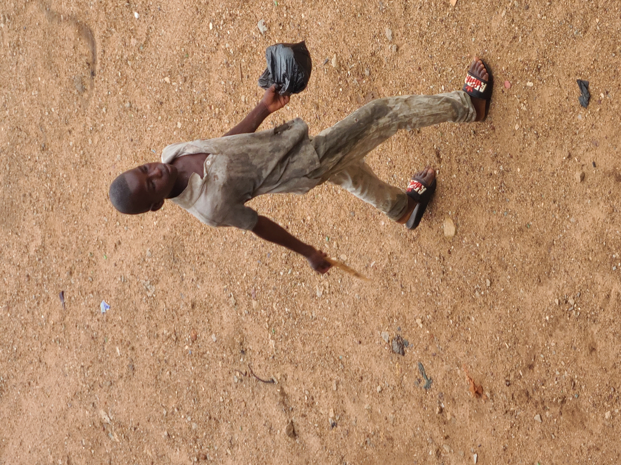 A young boy walks on a sandy surface in Gura Top, Nigeria. He is wearing a dirty, light-colored short-sleeved shirt and pants, and sandals. He is carrying a black plastic bag in his right hand and a stick in his left. The ground is covered with sand and small pebbles.