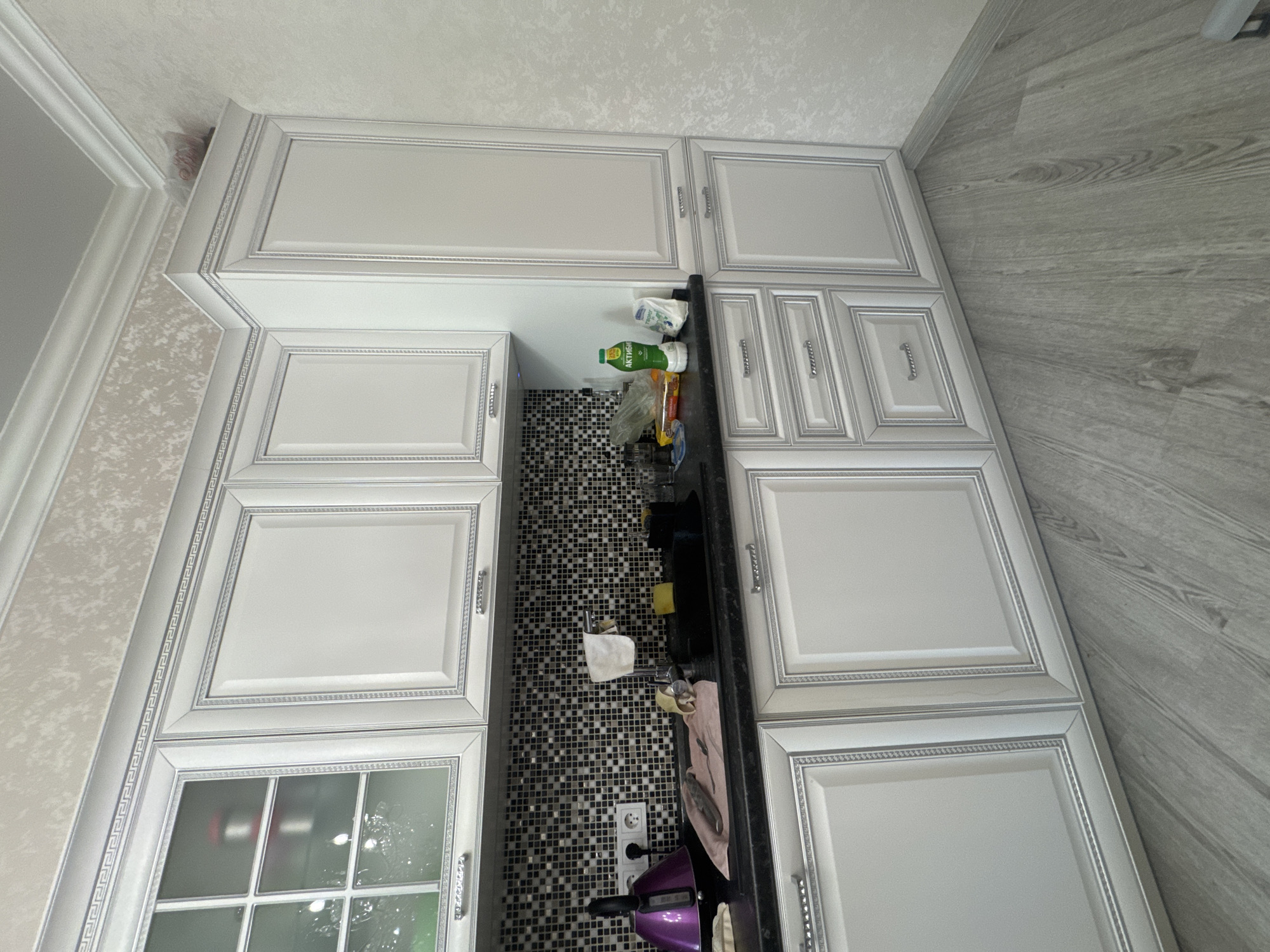 The image displays a high-angle view of a modern kitchen interior in Yuzhny, Russia, rotated 90 degrees clockwise. The kitchen features an L-shaped layout with light gray or off-white cabinetry, extending to the ceiling. The cabinet doors are adorned with ornate raised panels and decorative silver trim, incorporating a Greek key pattern, complemented by silver-toned handles. One of the lower cabinets on the left side of the frame has a glass-paneled door.
The walls are covered with a light, textured wallpaper, bordered by white crown molding at the ceiling. The floor, visible on the right, features light gray, wood-look laminate or tile planks.
A dark countertop runs along the base of the cabinets. A striking black and white mosaic tile backsplash fills the area between the upper and lower cabinets, particularly visible around the sink section. On the countertop, several items are arranged, including a green bottle labeled "АКТИБИО", various plastic bags, a sponge, and a pinkish cloth. A white electrical outlet with multiple plugs is situated above the countertop, connected to a purple small appliance, possibly a blender or kettle.
No people are visible in the scene, and no specific activity is occurring, presenting a static snapshot of the kitchen space. The lighting indicates it is daytime.