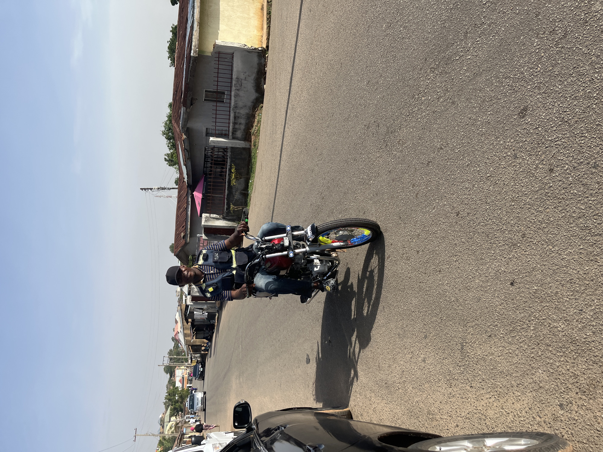 The image shows a man riding a motorcycle on a paved road in Jos, Nigeria. The man is wearing a striped shirt, jeans, a cap, and a vest. He is riding a red and white motorcycle with colorful decorations on the front wheel. The road is lined with buildings and parked cars. The sky is clear and blue. The location appears to be a residential area.