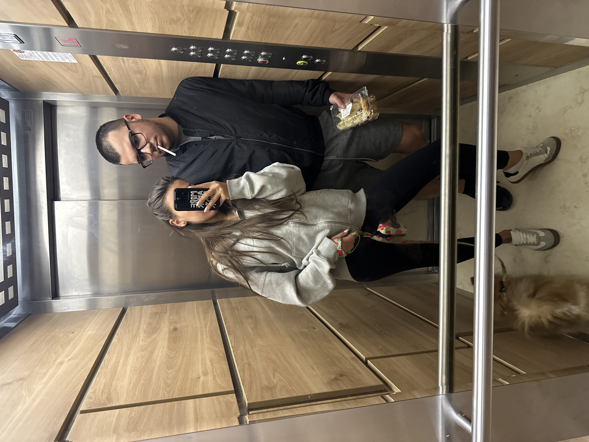 In an elevator with wood-paneled walls, a young man and woman are taking a mirror selfie. The man, wearing glasses and a black jacket, has a cigarette in his mouth. He holds a clear plastic container filled with food in his right hand. The woman, wearing a gray hoodie and black leggings, holds a smartphone in her left hand, which is angled to capture the reflection. Her right hand is holding a colorful leash. A small, fluffy dog with light brown fur is visible at the bottom of the elevator, reflected in the floor. The elevator interior appears modern and clean. The scene is likely in Sofia, Bulgaria.