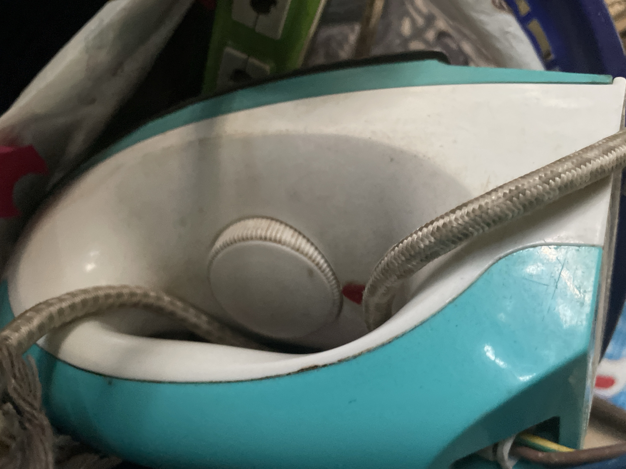 The image shows a close-up, overhead view of a turquoise and white steam iron. The iron has a textured, braided cord running across its surface. A dial with markings is visible on the top of the iron, along with a small red indicator light. The iron appears to be slightly dusty. In the background, a green power strip with several outlets is partially visible, along with other indistinct objects. The scene appears to be indoors, possibly in a home or workshop. There are no people visible in the image. The location of Jalingo, Nigeria is not depicted or suggested by any elements within the image itself.