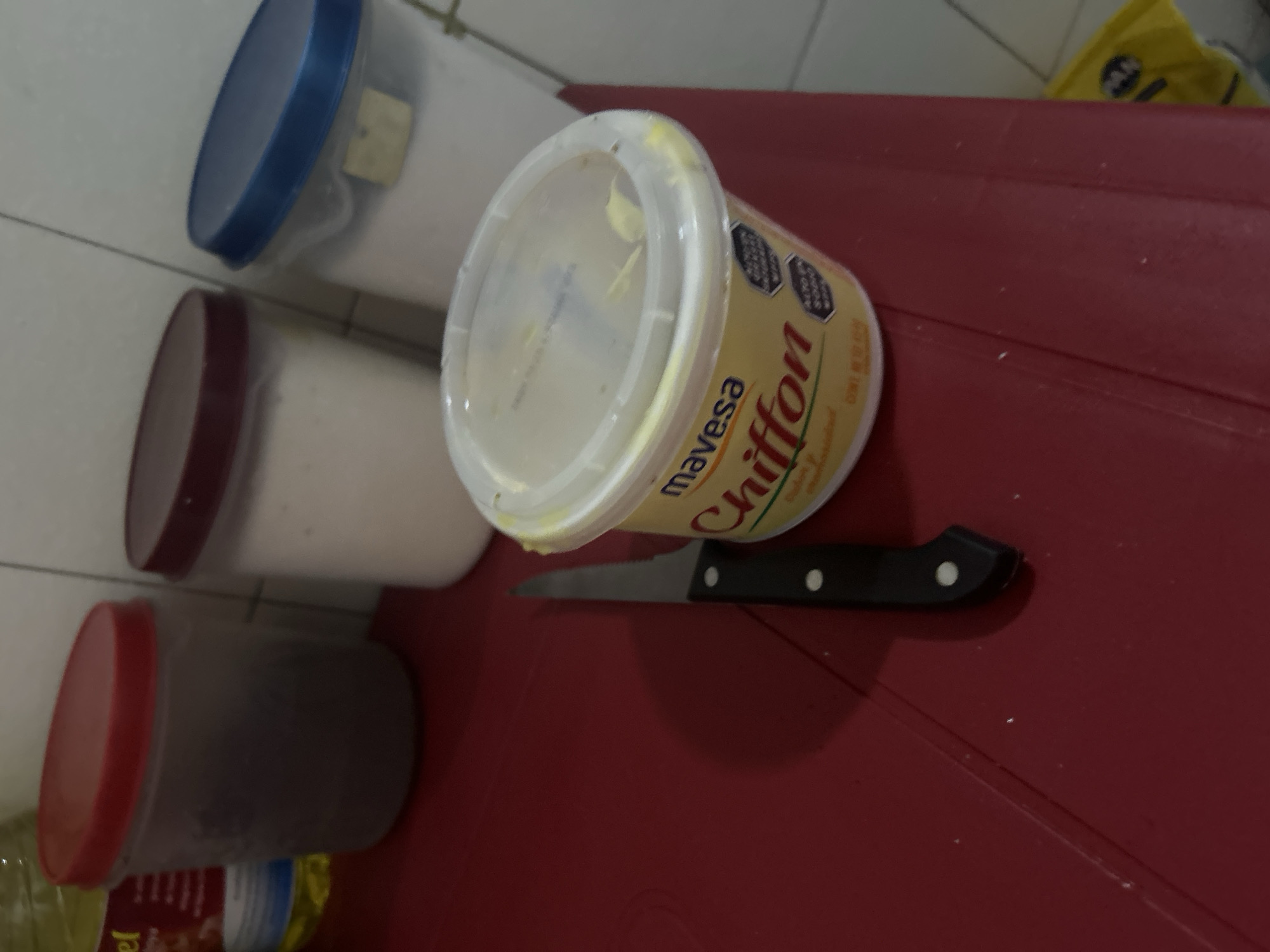 The image shows a red table with three plastic containers with lids of different colors (blue, red, and maroon), a container of Mavesa Chiffon margarine with a white lid, and a knife with a black handle. The location, San Juan de Los Morros, Venezuela, cannot be confirmed from the image.
