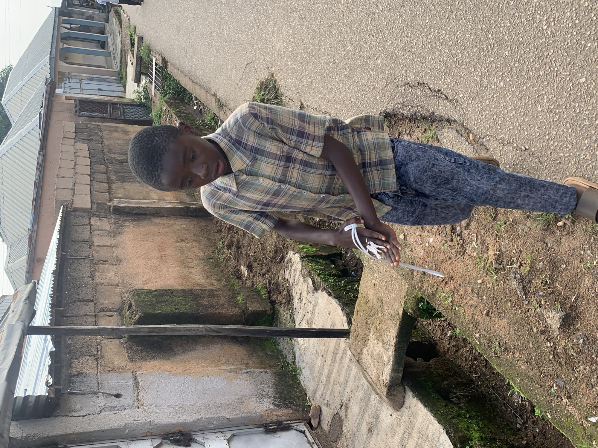 A young Black male wearing a plaid short-sleeved button-down shirt and distressed denim jeans walks on a paved road in Jos, Nigeria. He is holding a set of keys attached to a white wristband in his right hand. The road runs alongside a ditch filled with dirt and some vegetation. Behind the ditch, there is a concrete wall with a corrugated metal roof extension, and beyond that, residential buildings with metal roofs. The sky is overcast.