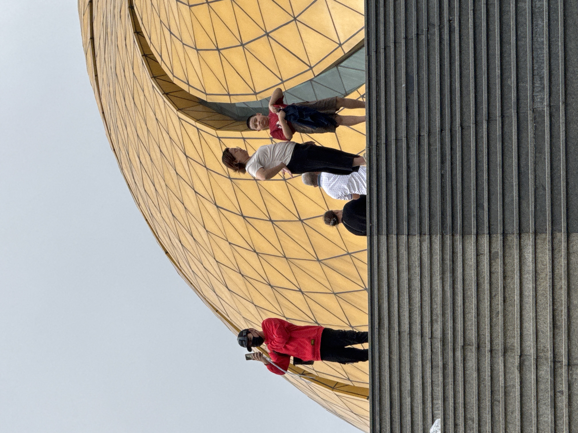 The image shows a group of people standing on a wide, grey stone staircase in front of a large, golden, geodesic dome structure. The dome is composed of numerous hexagonal panels and appears to be a modern architectural landmark. The sky is overcast, suggesting a cloudy day.
On the stairs, a young man in a red hoodie and black pants is standing on the left, wearing a black face mask and a black baseball cap. He is holding a smartphone up as if taking a photograph or video. To his right, further up the stairs, a woman with short brown hair is standing, wearing a light-colored t-shirt and black pants. She has her hands on her hips and is looking slightly to the left. Beside her, a young boy in a red t-shirt and khaki shorts is standing, holding a dark blue garment. Behind them, further up and to the left, two individuals are seated, their backs to the camera. One is wearing a white shirt, and the other a dark top.
The architectural style of the dome, with its distinctive golden geodesic pattern, is characteristic of the Lam Vien Square in Da Lat, Vietnam. Specifically, this dome is known as the "Flower Garden" or "Dalat Flower Park" pavilion. The structure is a prominent feature of the square, which is a popular tourist destination. The staircase appears to lead up to the entrance or a viewing area of this architectural marvel.