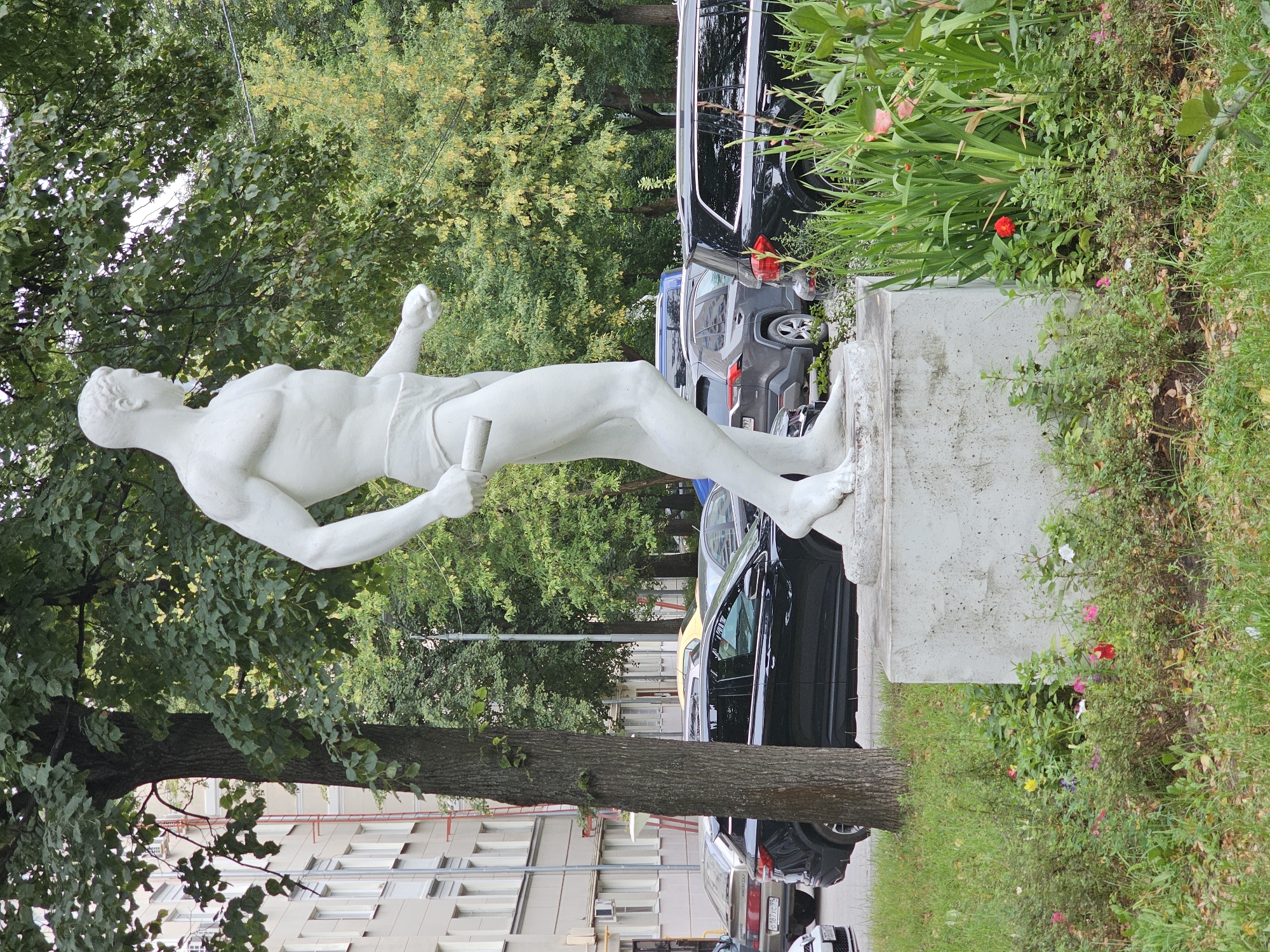 A white plaster statue of a muscular male athlete in a discus thrower's pose stands on a concrete pedestal. The statue faces right, with its left arm bent and holding a discus, and its right arm extended slightly forward. The athlete wears briefs. Behind the statue, a large tree with green leaves partially obstructs the view of a multi-story building with cream-colored walls and numerous windows. Several cars are parked on a street to the left of the building, including a black SUV and a darker vehicle. To the right of the statue, a dense green area with tall grass and red and pink flowers is visible, bordering a black SUV parked in the foreground. The overall scene appears to be an urban park or public space in Khimki, Russia.