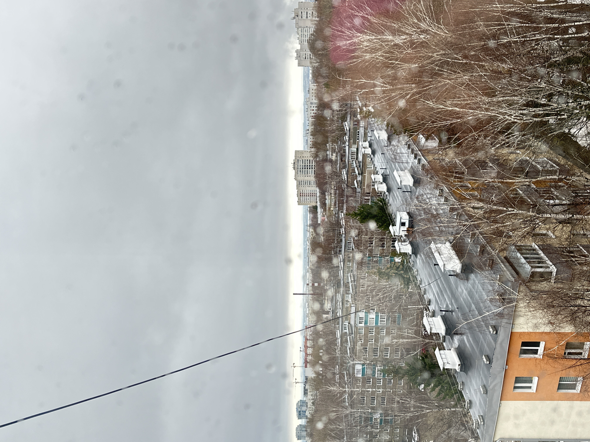 The image shows an overcast day in a city, likely Kazan, Russia. The view is from an elevated position, possibly a window or balcony, overlooking a street lined with multi-story apartment buildings, bare trees, and parked vehicles. The street is wet, possibly from recent rain or snow. A prominent power or communication line cuts across the foreground. The sky is a uniform gray, indicating a gloomy or inclement weather. There are a few buildings of the same architectural style, which are commonly found in Russian cities.