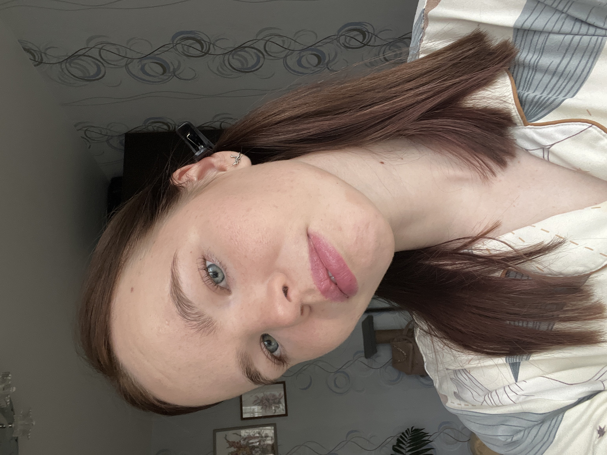The image, rotated 90 degrees clockwise, features a young woman posing for a close-up indoor portrait. She has long, straight, medium brown hair, some of which is pulled back with a black clip visible near her left ear. Her left ear also sports a small, intricate silver earring, possibly a helix piercing or cuff. She has light skin, blue-grey eyes, and well-defined eyebrows. Her lips are painted with pink lipstick, and her expression is calm and neutral, with a slight upward curve to her lips.
She is wearing a light-colored top with a subtle, abstract pattern, likely casual wear or pajamas, visible around her shoulders and chest.
The background consists of a wall adorned with a delicate, swirling grey and blue pattern. Below the wall on the left side of the frame, parts of two framed pictures with ornate designs and a green plant in a pot are visible. On the right side, a section of patterned bedding, possibly a duvet cover or pillow, can be seen. The lighting appears soft and even, suggesting an indoor setting, likely a bedroom or living space in Chaikovsky, Russia. There is no visible text in the image.