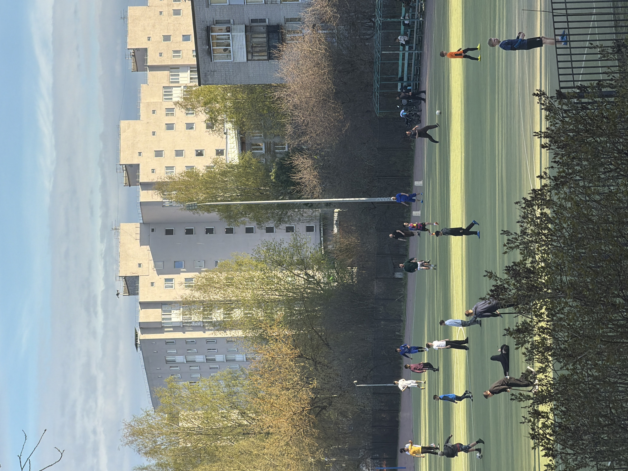 A green artificial turf soccer field is flanked by apartment buildings, trees, and chain-link fences. Several people are distributed across the field, some standing and walking, others lying down. A person in an orange shirt and a person in a blue shirt are running on the far side of the field, and another is kicking a soccer ball. The buildings have many windows and appear to be made of concrete. The trees are mostly bare, with some showing new leaves. The sky is blue with some wispy clouds. This scene is located in Myaglovo, Russia.