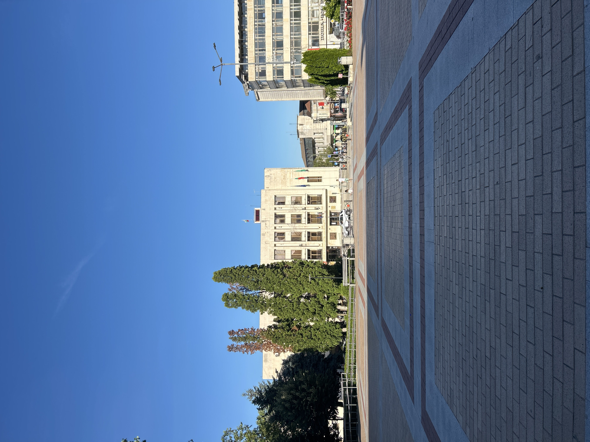 An outdoor, eye-level, wide shot shows a paved city square with a large, light-colored building in the background. The sky is clear and blue. The square is paved with light-gray bricks in a geometric pattern. A sidewalk with a decorative border separates the square from a grassy area with several tall, slender evergreen trees and one tree with reddish-brown foliage. Behind the trees is a large, rectangular building with a facade of light stone or concrete, featuring multiple floors of windows and a central protruding section with a flat roof. Two flags, one red and one green, are visible on the building's facade, along with a white flag at the top. To the right of this building, a taller, modern building with a light-colored facade and many windows stands. A street lamp with multiple lights is positioned in front of this building. In the mid-ground, several people are walking and standing. A white car is parked in front of the stone building. The scene appears to be a public plaza in front of municipal buildings on a clear day.