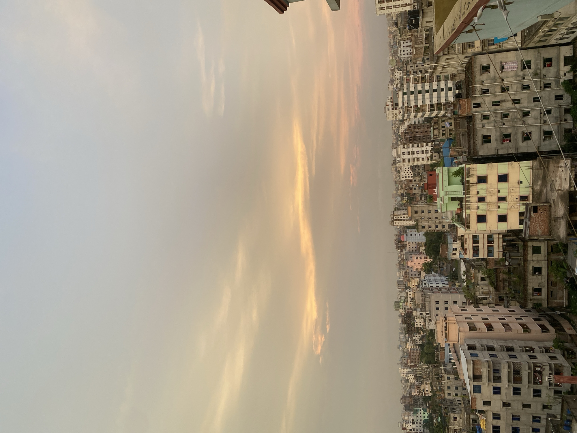 This image captures a dense urban landscape of Dhaka, Bangladesh, under a hazy sky at what appears to be dusk. The scene is dominated by numerous multi-story buildings packed closely together, characteristic of the city's high-density development. The buildings vary in color, from weathered concrete to pastel shades, and many show signs of age and ongoing construction or disrepair. Rooftops are cluttered with water tanks, satellite dishes, and various makeshift structures. Vegetation is sparsely visible, primarily as small trees or plants growing on balconies and rooftops. The sky is a soft gradient of pale yellow, orange, and pink, suggesting the last light of the sun breaking through the atmospheric haze. There are no visible individuals in the frame, and the overall impression is one of a bustling, densely populated urban environment.