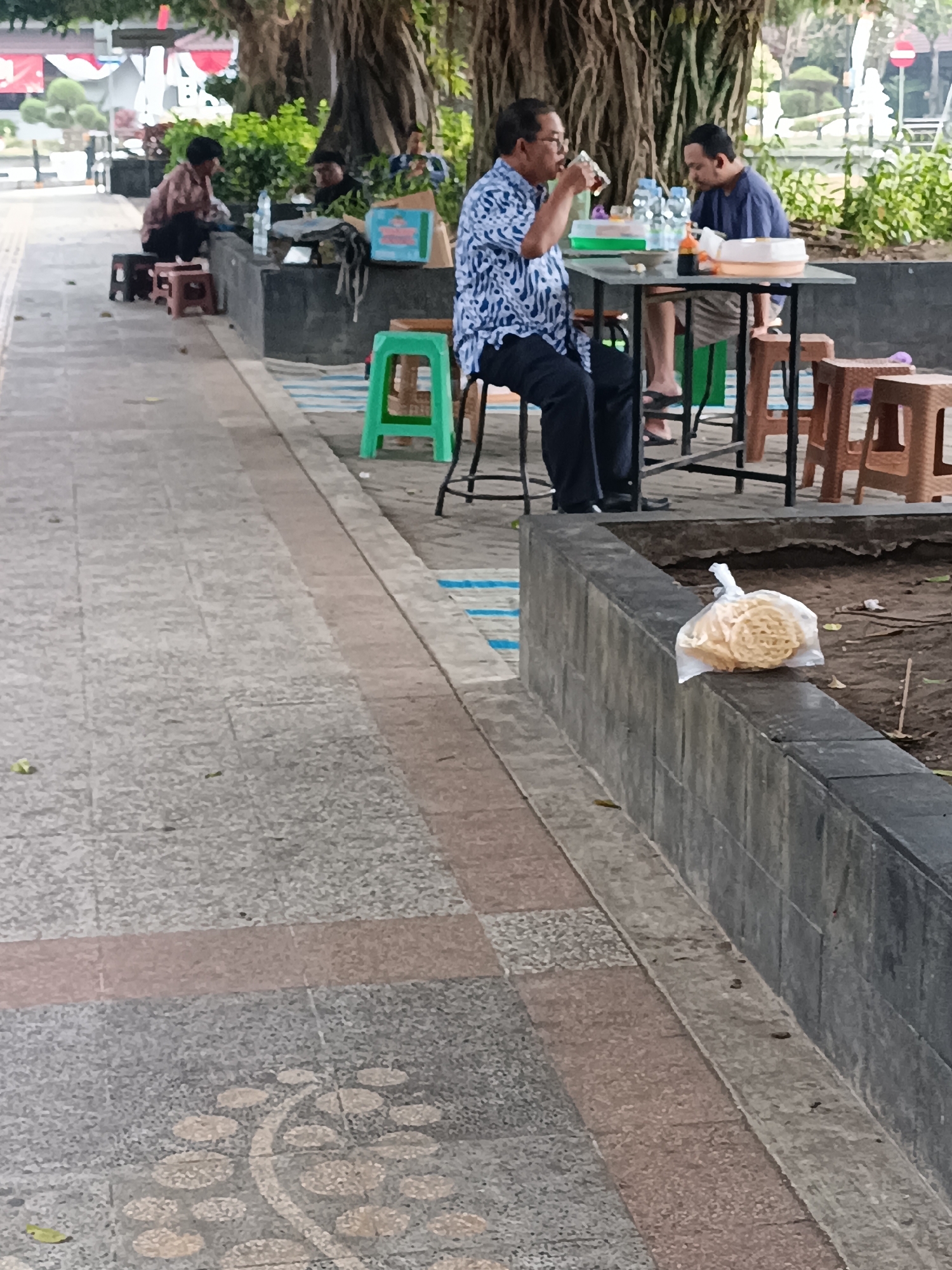 A street scene in Sleman, Indonesia, features two men seated at a table outdoors. One man, wearing a blue and white patterned shirt and dark pants, is drinking from a glass. The other man, in a blue shirt and striped shorts, is looking down. They are seated at a square table with several bottles of water and a dish of food. Behind them, a large tree provides shade, and lush green foliage is visible. In the foreground, a textured paved walkway runs alongside a raised concrete planter. A clear plastic bag containing circular crackers rests on the edge of the planter. Several small plastic stools are also scattered around the area. Other individuals are visible in the background, further down the walkway, engaged in their own activities.