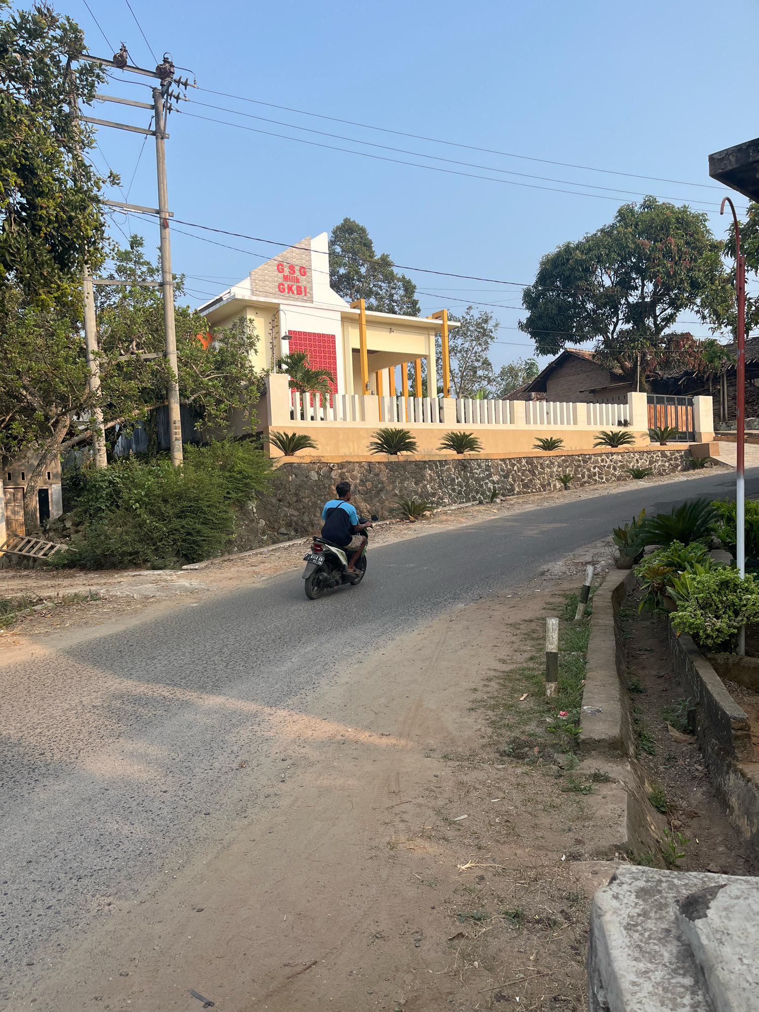 A person rides a motorcycle on a paved road in Margodadi, Indonesia. The person is wearing a blue shirt and dark shorts, and is carrying a dark backpack. The motorcycle is black and white. The road is lined with trees and bushes on both sides. In the background, there is a building with a white and yellow facade and a red patterned section. There is also a stone wall with plants in front of the building. A power pole with wires stands to the left of the road. The sky is clear and blue.