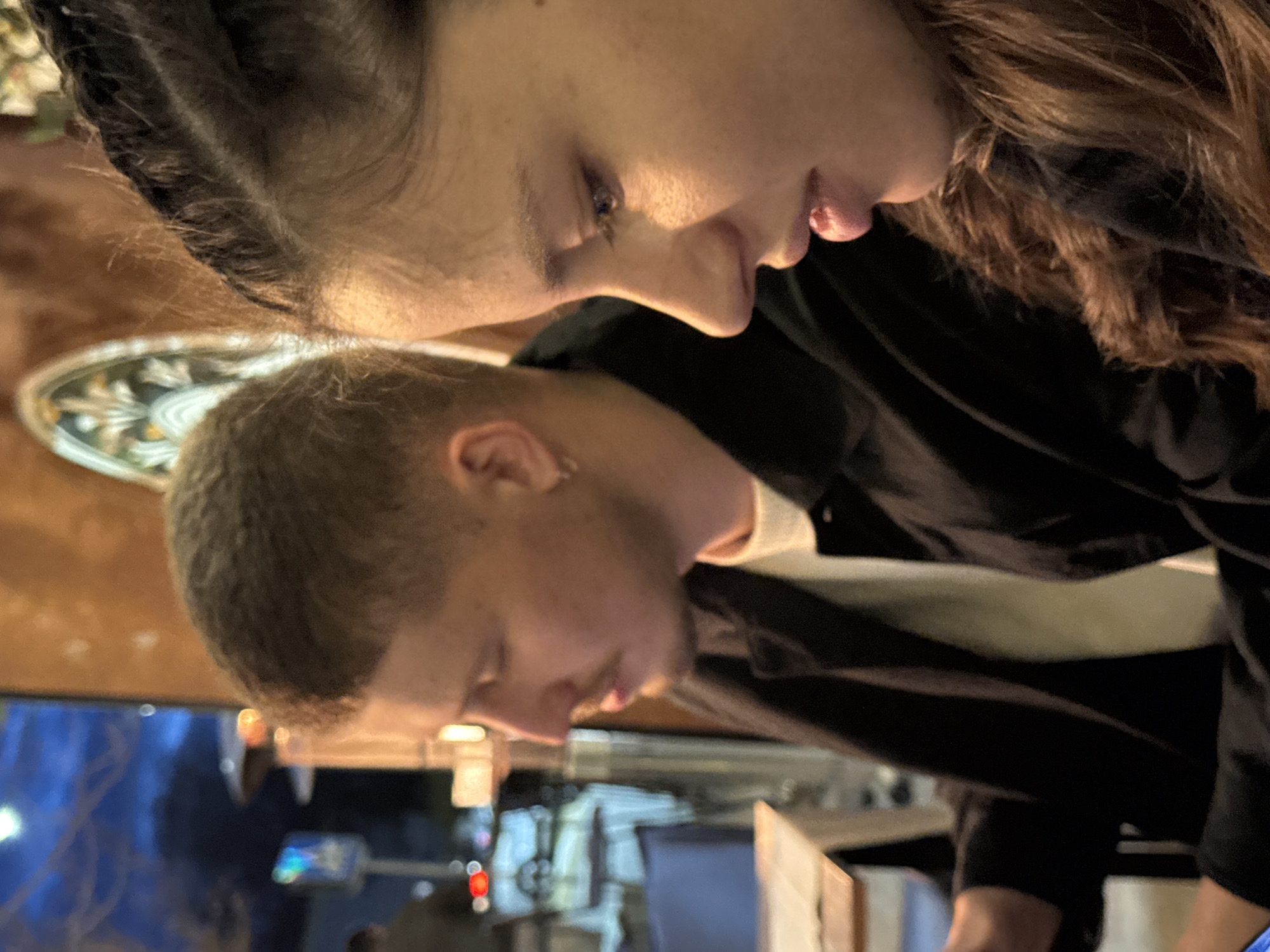 This image captures two individuals seated within a commercial establishment, likely a restaurant or cafe, during nighttime in the city of Unknown, Unknown.
The female subject, positioned on the right foreground, is shown in profile, looking downwards with her mouth slightly open. She has fair skin, dark brown hair styled with visible braids or twists near the crown, and wears a dark top. The male subject, positioned on the left midground, is also in profile, looking downwards. He has fair skin, short brown hair, a light mustache, and a silver hoop earring on his left ear. He wears a dark jacket over a light shirt.
The indoor setting features warm ambient lighting and a decorative, patterned wall art piece displaying greens, yellows, and browns. A light-toned table and a dark blue chair are partially visible. Through a window behind the subjects, a blurred exterior urban environment is visible under a dark blue sky, illuminated by streetlights, red and white vehicle lights, and an active outdoor patio heater. A blue informational sign is also discernible outside.