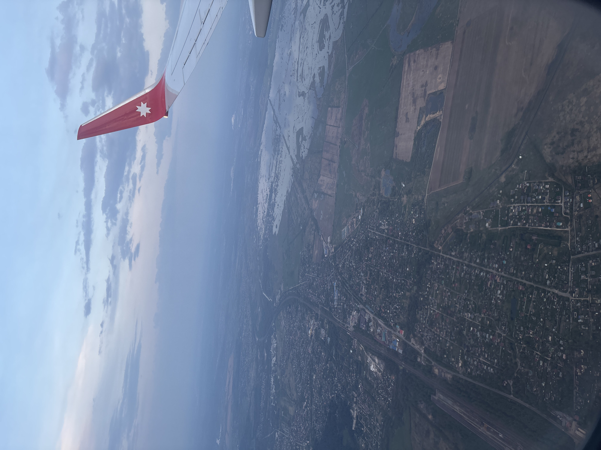 The image depicts a view from an airplane window, showing part of an airplane wing in the upper left corner. The wing is red with a white, eight-pointed star. Below the wing, the landscape is visible. This area appears to be a mix of agricultural fields, bodies of water (possibly flooded areas or rivers), and residential zones. The residential areas consist of dense clusters of buildings and houses, typical of a city's suburbs. In the lower portion of the image, there is a visible road and railway tracks. Based on the wing's identification mark and the general landscape features, the location is likely near the city of Domodedovo, Russia, as it is known to host Domodedovo International Airport and features similar terrain. There is no visible evidence of people or specific actions in the image.