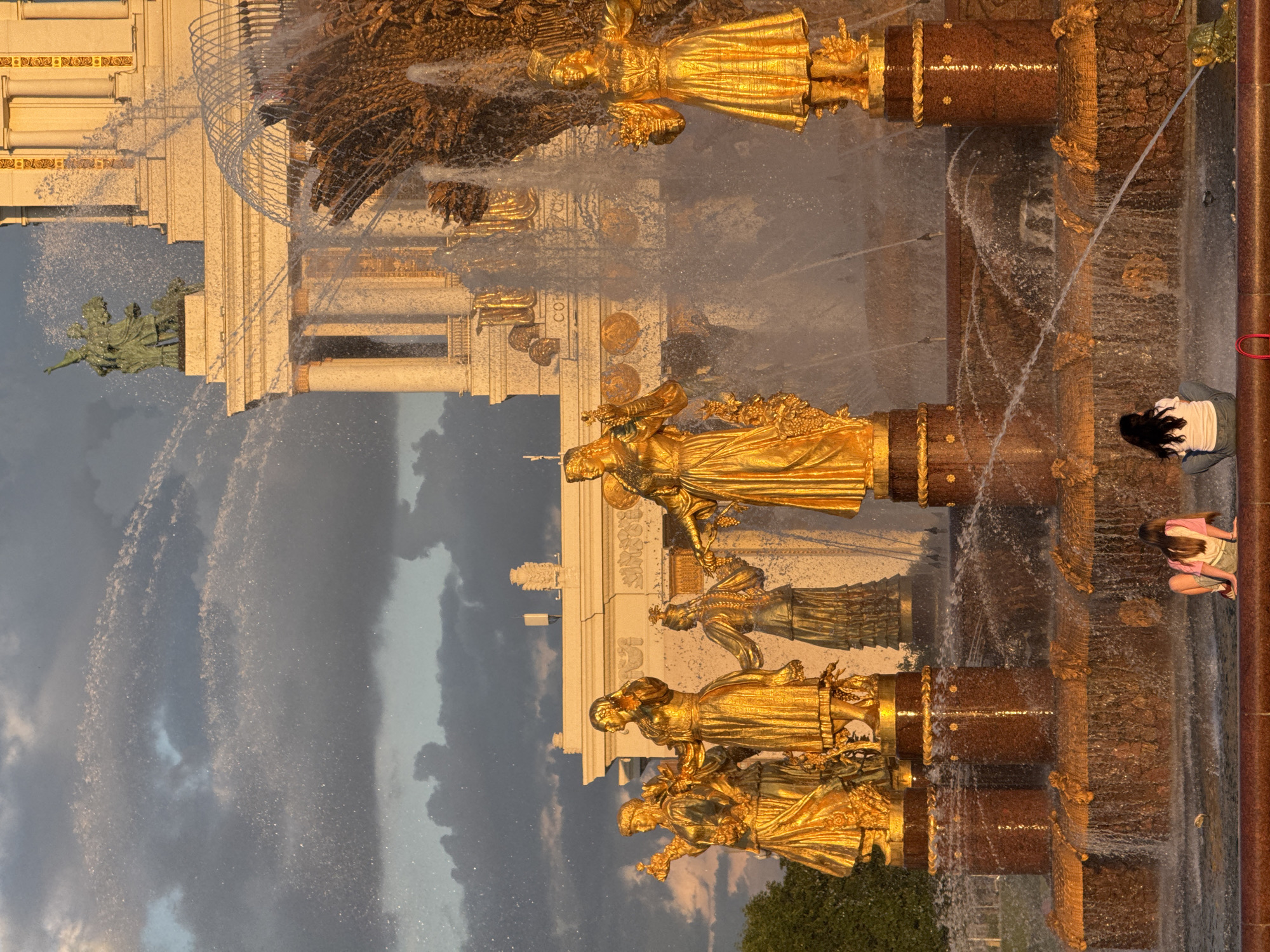 A grand, multi-tiered fountain, likely the "Friendship of Peoples" Fountain at VDNH in Moscow, dominates the scene under a dramatic, cloudy sky. The fountain is adorned with numerous large, gilded statues, predominantly female figures, rendered in a striking golden hue. These statues stand on pedestals and platforms, some holding symbolic items like sheaves of wheat or garlands. Water sprays dynamically from various points, creating shimmering jets and cascades that catch the warm, late afternoon or early evening light.
The fountain's base and supporting structures are constructed from a rich, reddish-brown stone, possibly granite, featuring intricate golden decorative elements and carvings. On the right side of the fountain, two individuals are visible. One person with long dark hair is seated or kneeling by the fountain's edge, wearing a white top and blue bottoms, seemingly observing the water. Another person, partially visible further to the right, wears a pink top and light-colored shorts.
In the background, a monumental, light-colored building with classical architectural features, including columns and cornices, stands behind the fountain. To the left of this main structure, a smaller green-patinated statue of a figure on a pedestal is discernable. The sky presents a dramatic contrast with dark, heavy clouds interspersed with brighter patches, enhancing the atmospheric quality of the scene. The overall lighting bathes the golden statues and architecture in a soft, warm glow. No legible text is visible in the image.