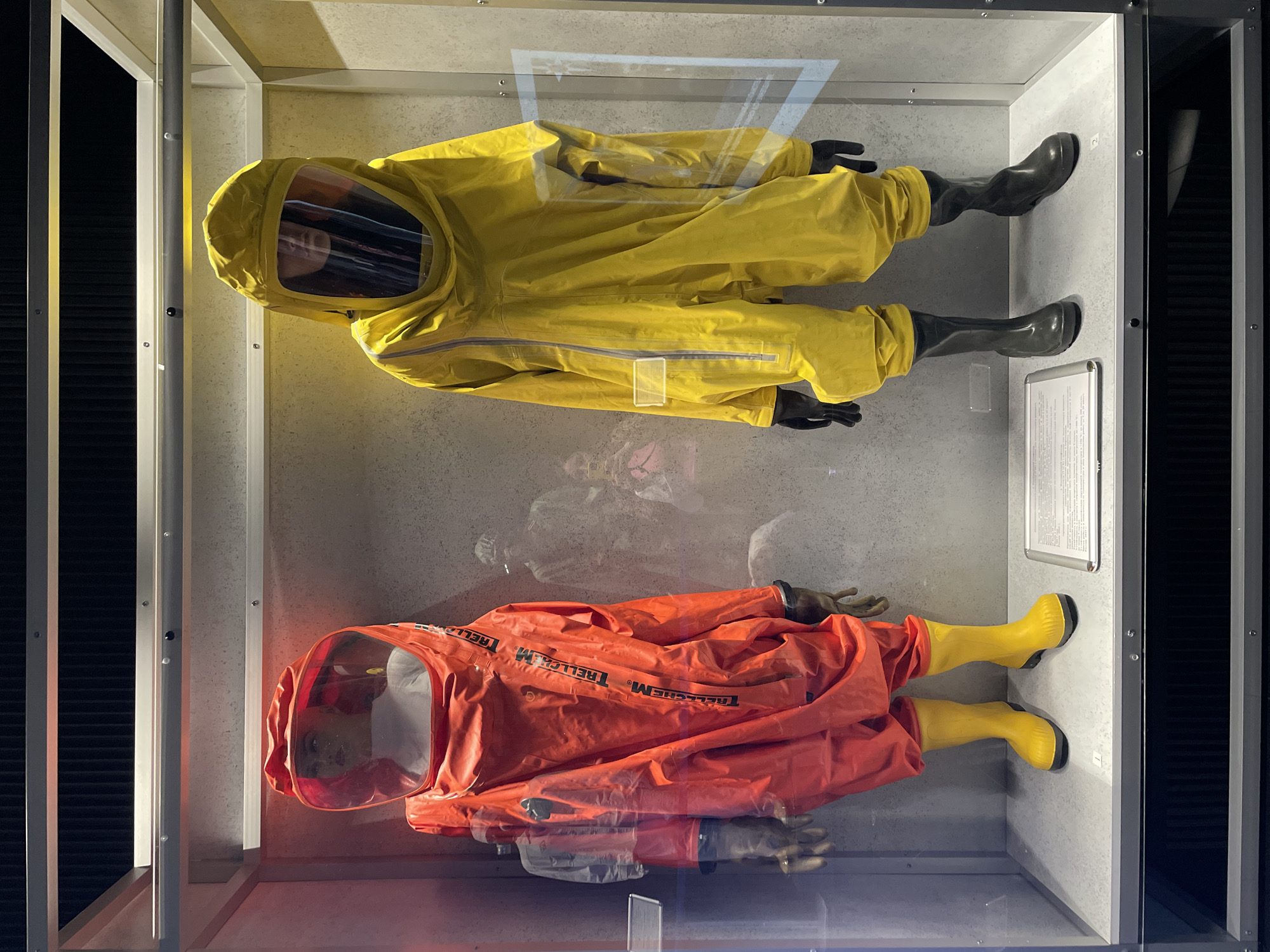 The image shows two hazmat suits displayed in a glass enclosure. The suits, positioned one above the other, appear to be on mannequins. The top suit is yellow with black gloves and black boots. The lower suit is red with the word "TRELLECHEM" printed on it, and has tan gloves and yellow boots. A sign is visible to the right of the yellow suit. The enclosure is framed with white and black material. The image was taken at the Museum of Cosmonautics in Moscow, Russia.