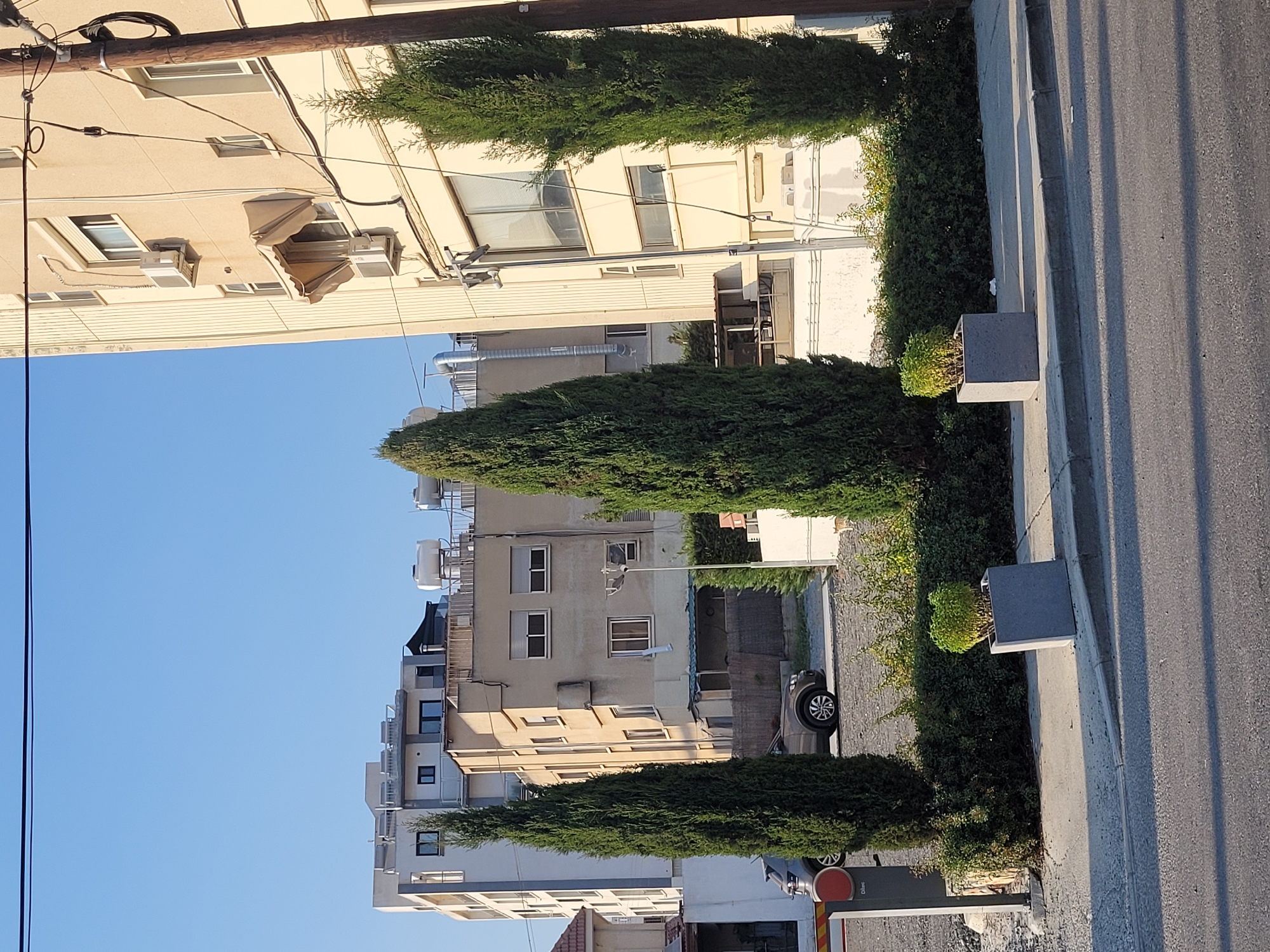 The image depicts a street scene in Limassol, Cyprus, on a clear day with a bright blue sky. In the foreground, a paved road leads into the frame, with a sidewalk bordering it. On the sidewalk, there are two large, square, grey planters, each containing a small green bush. Behind the planters, a dense, manicured hedge runs along a low wall.
Several buildings are visible. The closest building on the right is a multi-story structure with a light-colored facade, featuring multiple windows, some with awnings, and visible air conditioning units. A wooden utility pole with wires attached stands in front of this building. To the left, a narrower, lighter-colored multi-story building is visible, with rooftop water tanks and solar panels. Further back, another building with a beige facade and numerous windows is seen, with a dark slatted fence in front of its lower level. Two cars are parked on a gravel lot behind the hedge and in front of the buildings. Tall, slender cypress trees are strategically placed in front of the buildings, framing the scene. A red and grey automated gate, likely for a parking area, is partially visible on the far left. The overall impression is an urban environment with residential and commercial buildings, complemented by landscaping.