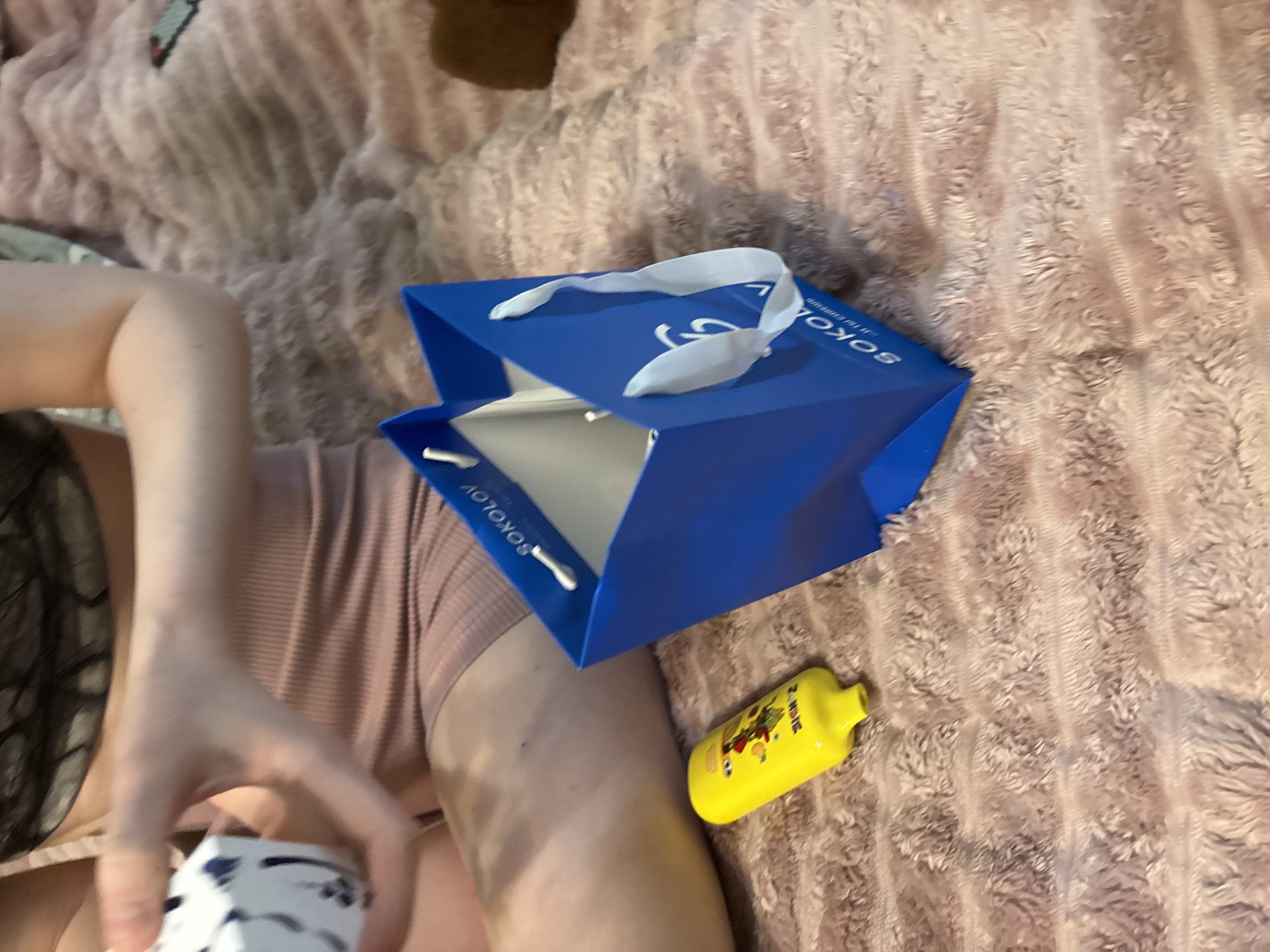 In Sinyavino, Russia, a person is holding a white object with blue designs. Nearby, a bright yellow bottle with cartoon characters and text is lying on a textured, light pink blanket. A blue gift bag with white ribbon handles and the word "SOKOLOV" printed on it is open and stands on the blanket. The person is wearing a light pink ribbed garment. The scene appears to be indoors, on a soft surface.