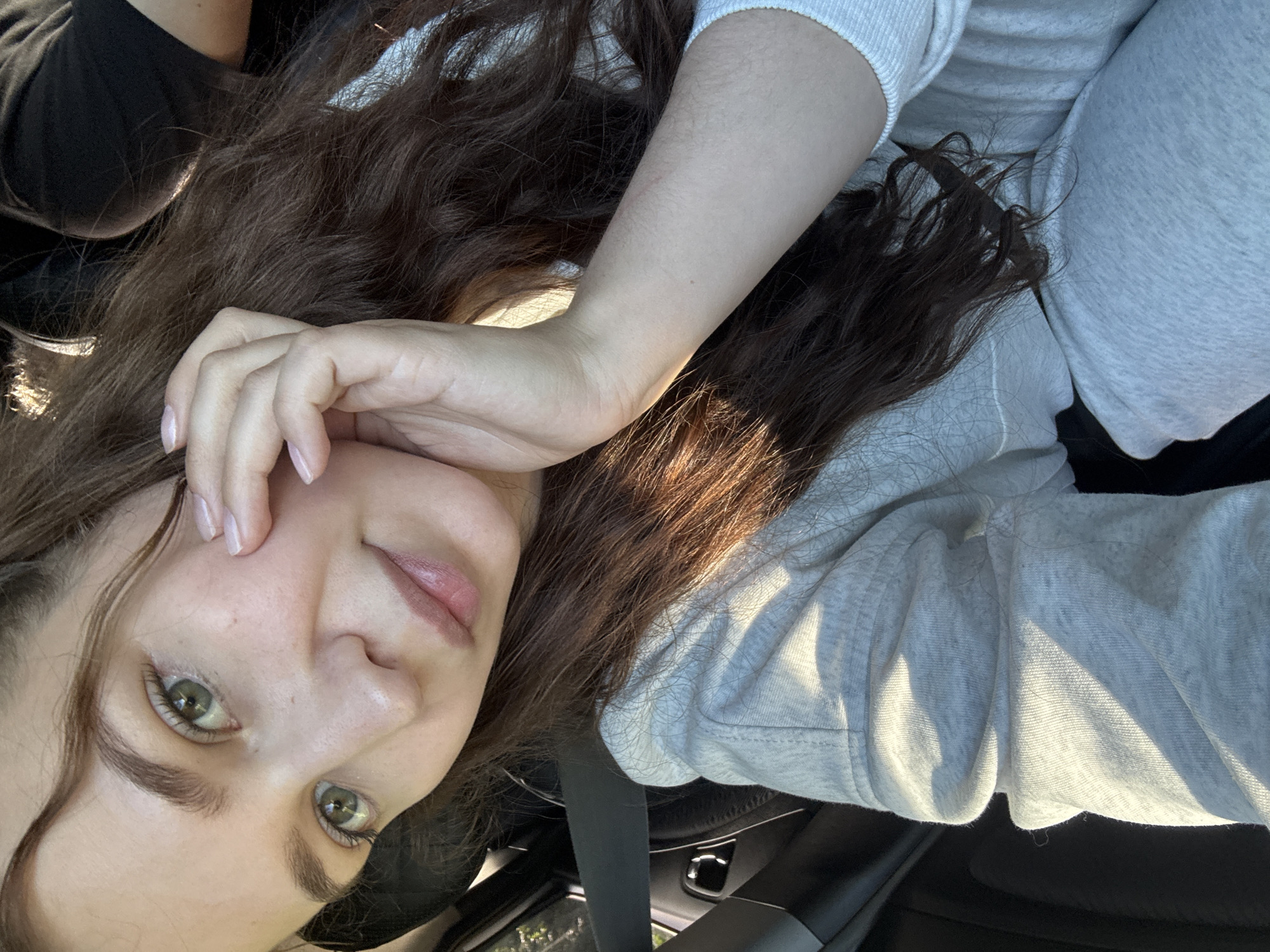 A young woman with long, wavy brown hair and green eyes is taking a selfie in a car. She is wearing a light grey hooded sweatshirt and has her right hand resting on her cheek. The car interior, including a seatbelt and part of the dashboard, is visible in the background. The lighting suggests it is daytime, with sunlight casting shadows. The provided image does not contain enough information to determine the specific location within Lukintsy, Russia.
