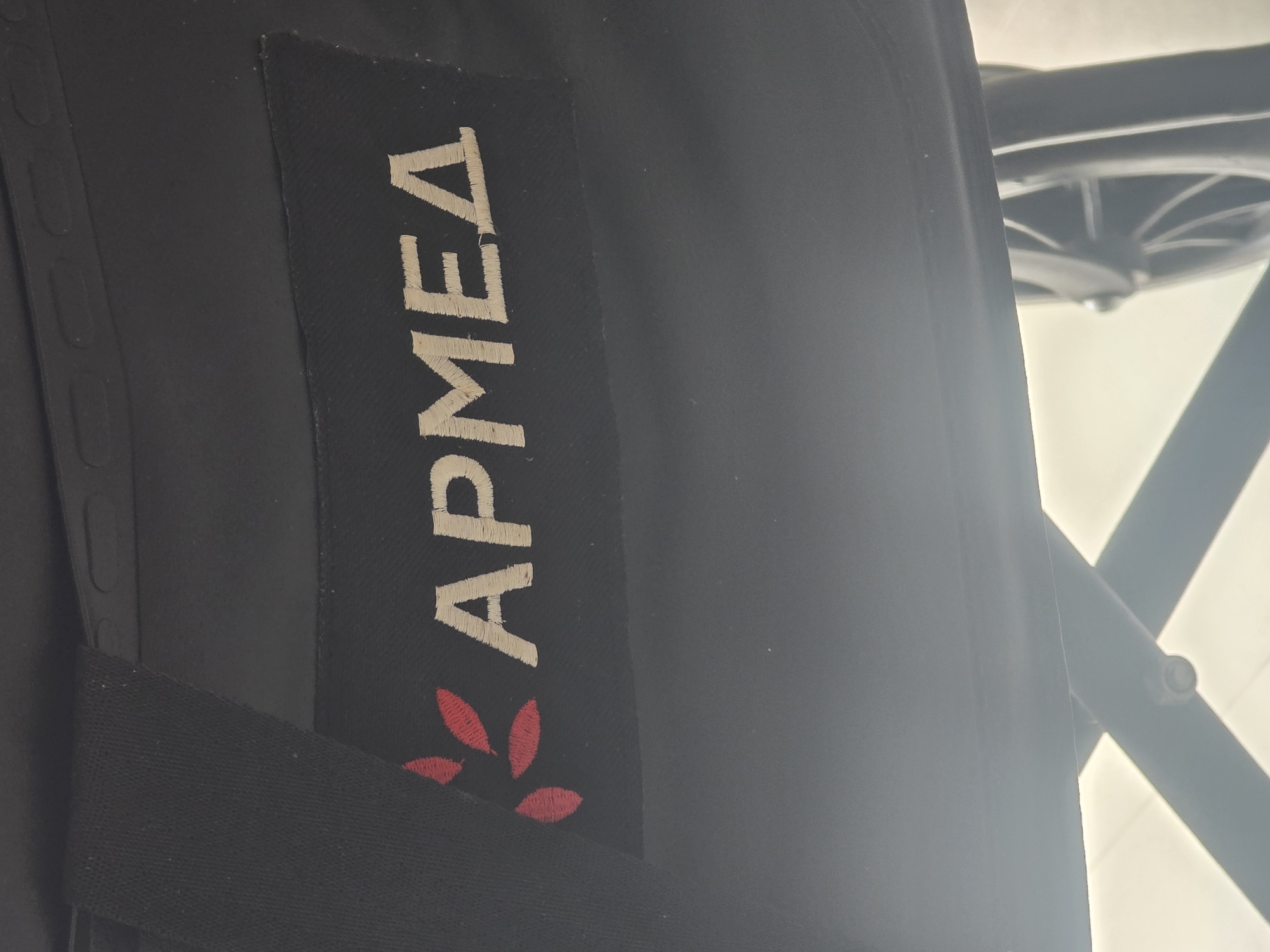 The image shows a close-up view of a black fabric patch with embroidered text and a logo. The text reads "ARMED" in a light beige color. To the left of the text is a stylized logo with four red petal-like shapes arranged in a circular pattern. The patch is affixed to a larger piece of black fabric, possibly from a bag or piece of equipment. The background is dark, with some out-of-focus elements suggesting a dimly lit environment. There are no people, specific landmarks, or details that would allow for the identification of a location in Ulan-Ude, Russia, or any other city. The image appears to be a detail shot of a branded item.