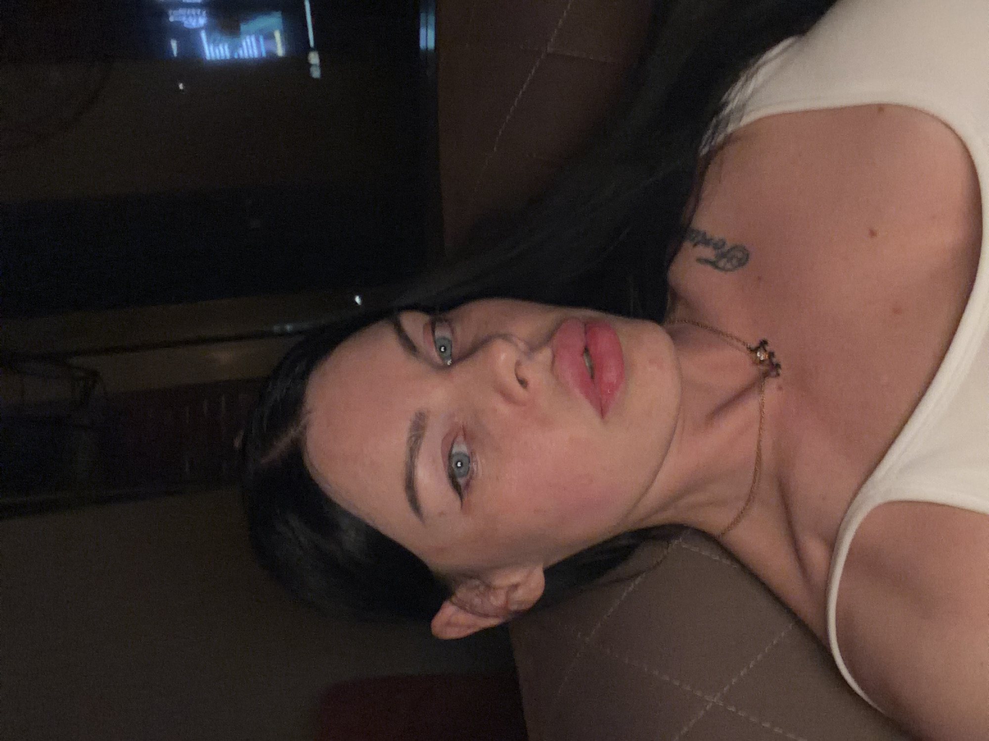 The image shows a close-up, selfie-style shot of a person with dark hair and blue eyes. The person is wearing a white top and a thin gold necklace with a charm. A tattoo is visible on the person's upper chest/shoulder area. The background is dark and appears to be indoors, possibly in a dimly lit room with a television screen visible in the upper right corner, displaying text. The lighting is soft, casting shadows on the person's face. The overall impression is of a casual, personal photograph taken in a private setting. There is no information in the image to suggest the location is the city of Ufa, Russia.