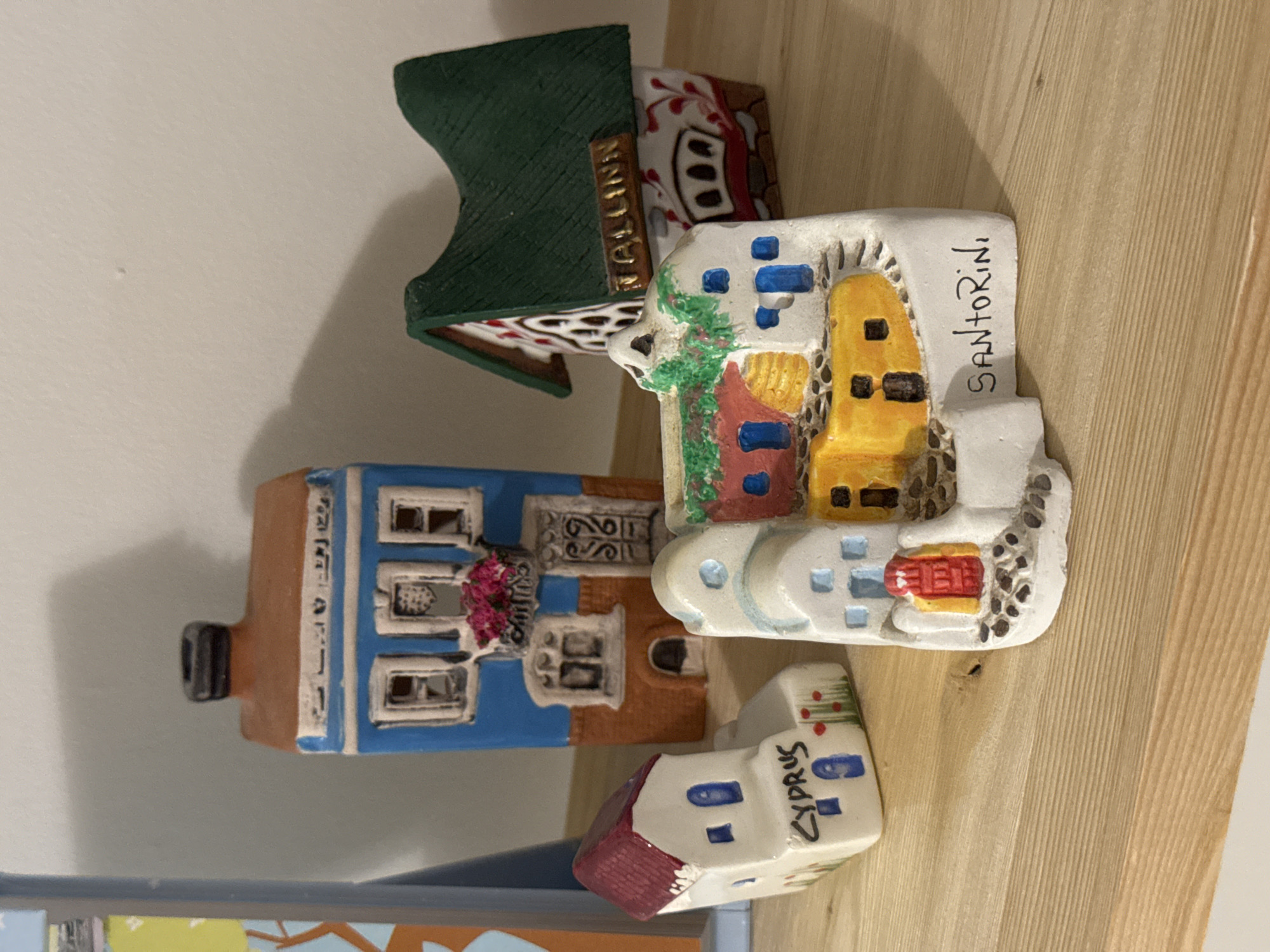 The image shows four miniature souvenir buildings. From left to right, they represent buildings from Tallinn, Santorini, a generic European city and Cyprus, respectively. The scene is set indoors on a wooden shelf. There is no indication of Izhevsk, Russia in the image.