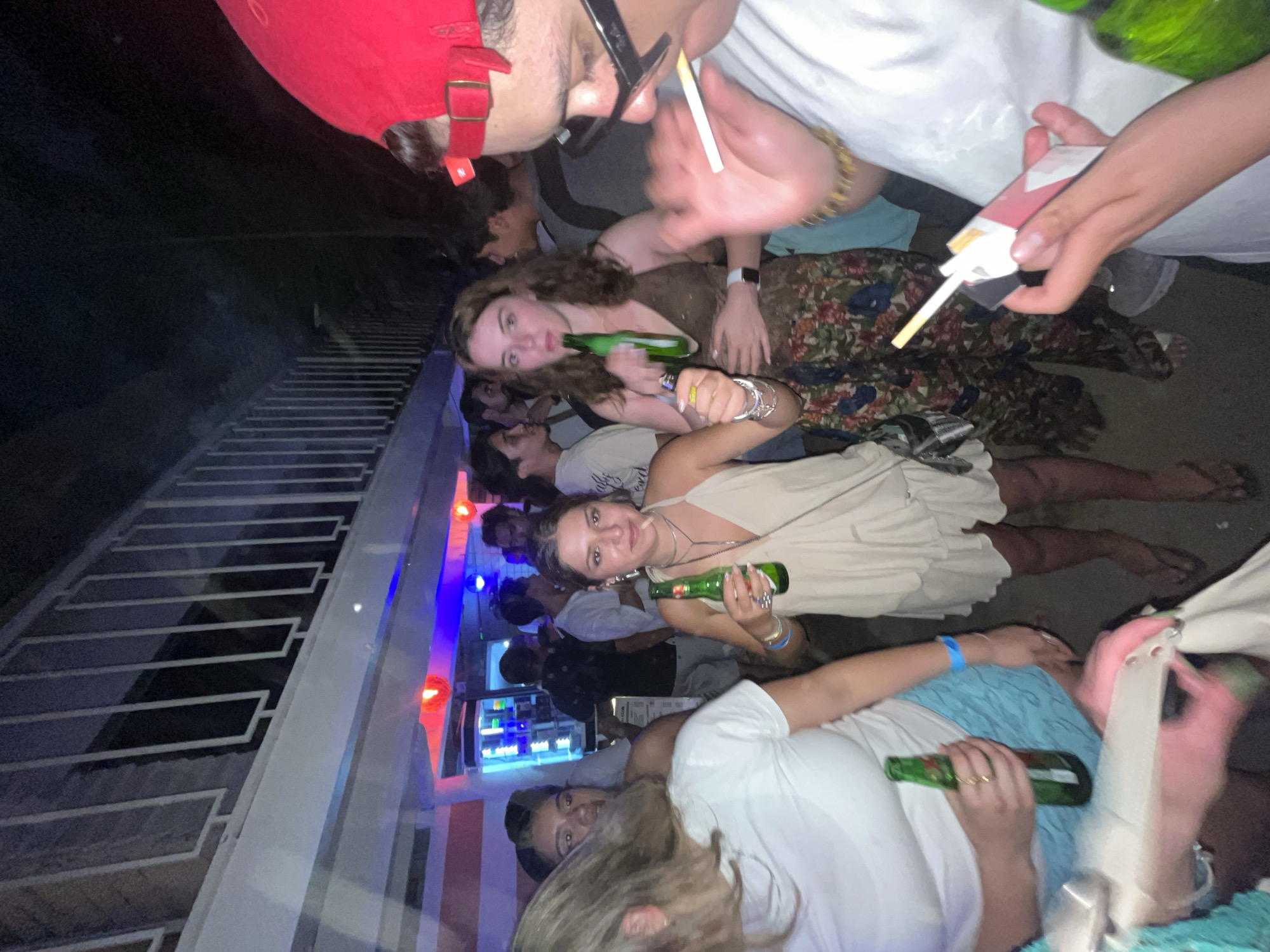 The image appears to depict a group of people at a gathering in Brisas de Zicatela, Mexico. Several individuals are holding green beer bottles. One person is holding a lit cigarette, while another holds a pack of Marlboro cigarettes. The setting seems to be an outdoor venue, as indicated by the presence of a white staircase with decorative metal railings illuminated by multi-colored lights. The overall atmosphere suggests a social event with a relaxed, informal vibe.