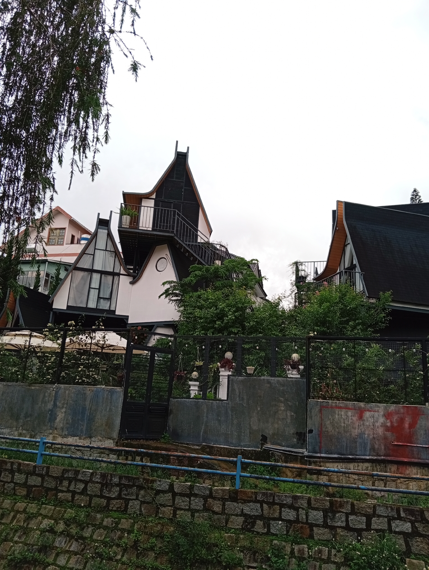 The image captures a unique cluster of buildings, likely a resort or hotel, set against an overcast sky in Xuân Hương - Đà Lạt, Vietnam. The scene is dominated by fantastical, whimsical architecture, characterized by steep, often curved or pointed, dark-colored roofs that contrast with predominantly light-colored (white or pale) facades.
In the foreground, a rough-hewn stone retaining wall slopes from the lower left, topped by a blue metal railing, some parts showing rust. Above this, a concrete wall with sections appearing weathered and some red discoloration supports dark metal gates, beyond which lush green bushes and flowering plants are visible.
The buildings display diverse designs:
* On the far left, a more traditional, pale pink building with a gabled roof and multi-paned windows is partially seen.
* The central and right structures are more striking, featuring dramatic rooflines, large geometric windows (some triangular), and external metal staircases leading to balconies. One balcony on the upper right has a potted plant. A distinctive circular window is visible on one white wall.
* Another building to the far right, largely obscured by trees, also features a dark, curved roof.
Lush green trees and dense shrubbery surround the properties, partially concealing lower levels and adding to the natural setting. Overhanging pine-like branches with dark green needles frame the top left corner of the image.
The weather appears cloudy with diffused light, but no specific time of day is indicated beyond daylight. No people or visible text are present. The overall impression is one of creative, unconventional architecture integrated into a verdant landscape.