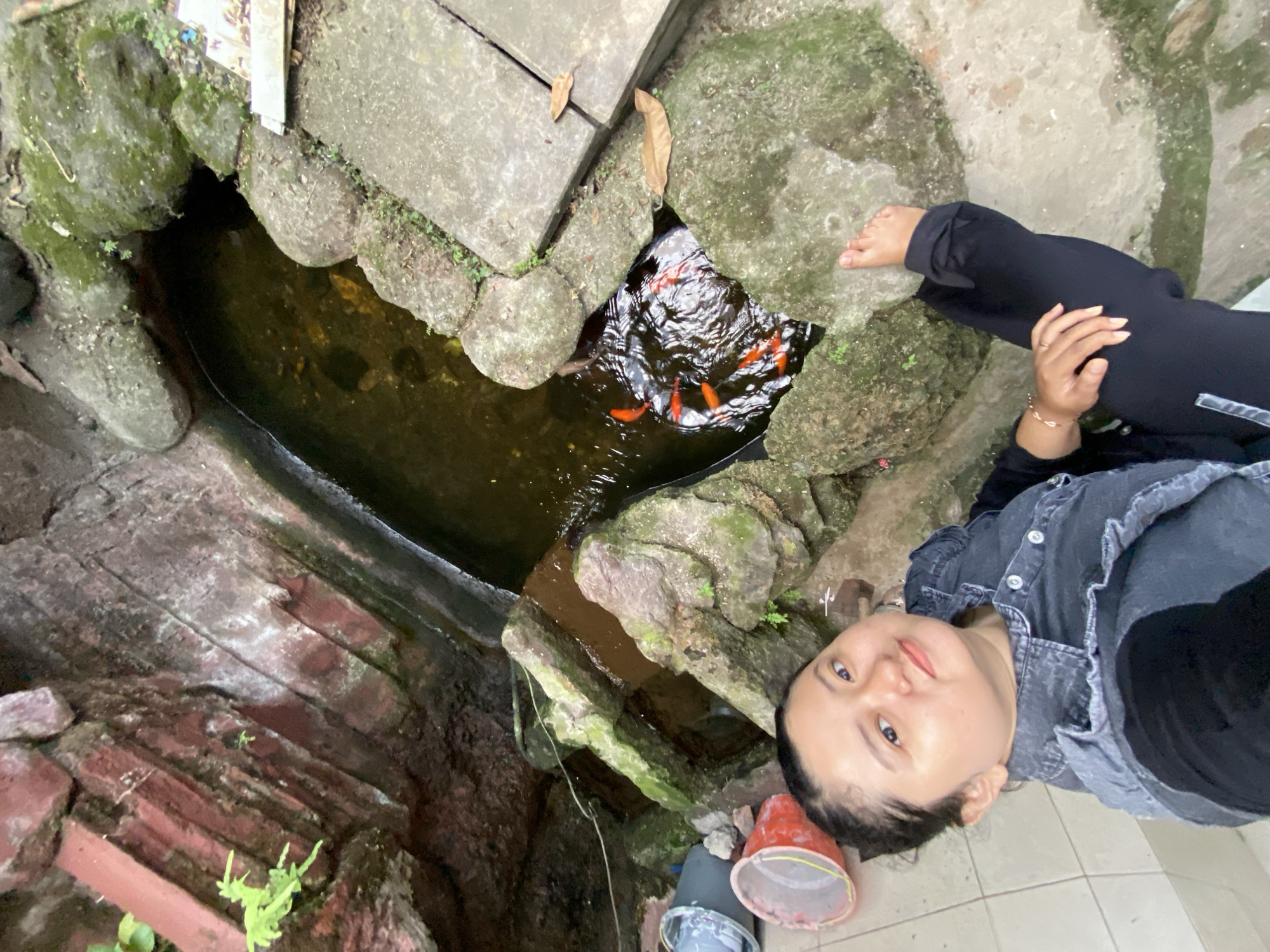 In Gelam, Indonesia, a young woman with dark hair pulled back is taking a selfie outdoors. She is wearing a dark denim vest over a black top and black pants. Her left leg is extended, and her right foot is visible near a rocky pond. The pond contains several orange goldfish swimming in dark water. The surrounding environment features moss-covered rocks, some red bricks, and a concrete walkway. Two buckets are visible on the left side of the image, one red and one gray, with some light-colored items inside the gray one. The scene appears to be a natural or semi-natural outdoor setting.