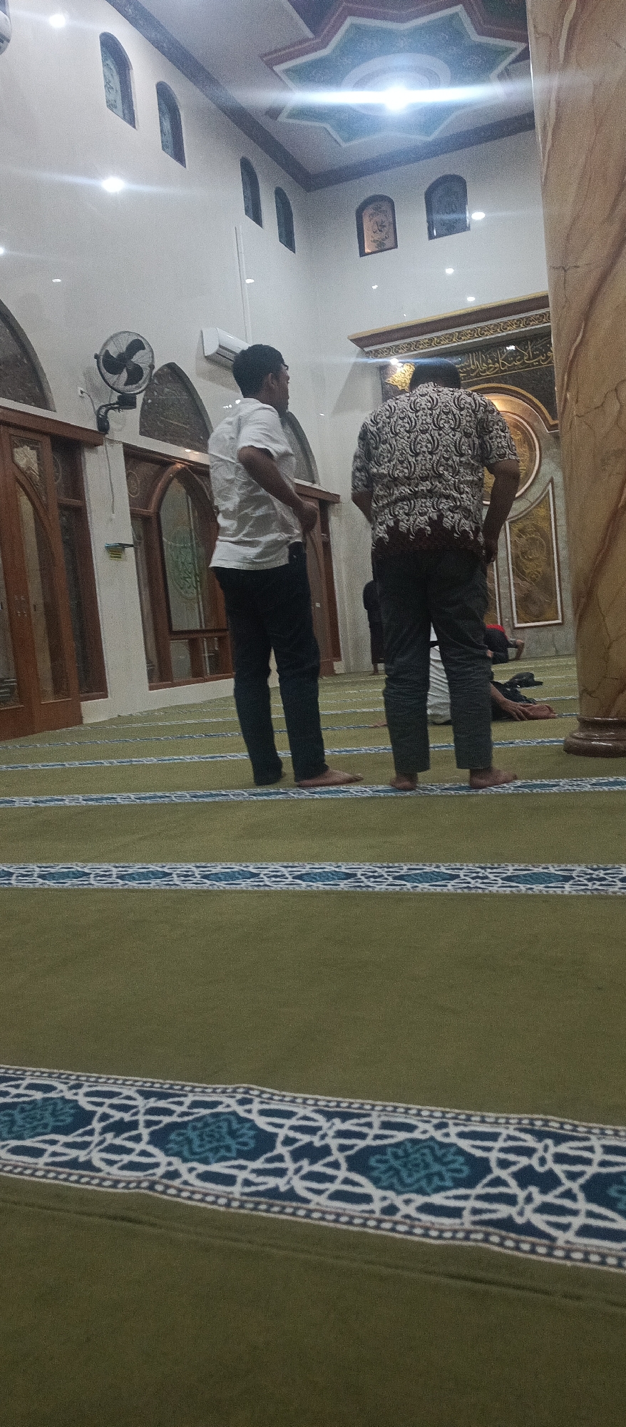 The image depicts the interior of a mosque in Arjasa, Indonesia. Two men are standing in the foreground, facing away from the camera. The man on the left is wearing a white short-sleeved shirt and dark trousers. The man on the right is wearing a patterned short-sleeved shirt and dark trousers. Both men are barefoot. In the background, several arched windows are visible along the wall, and ornate calligraphy is displayed on the wall above. A ceiling fan is mounted on the wall to the left. The floor is covered with a green carpet with blue and white patterned borders. Another person is visible lying down on the carpet in the middle ground. The architecture suggests a traditional Islamic design with ornate decorative elements.