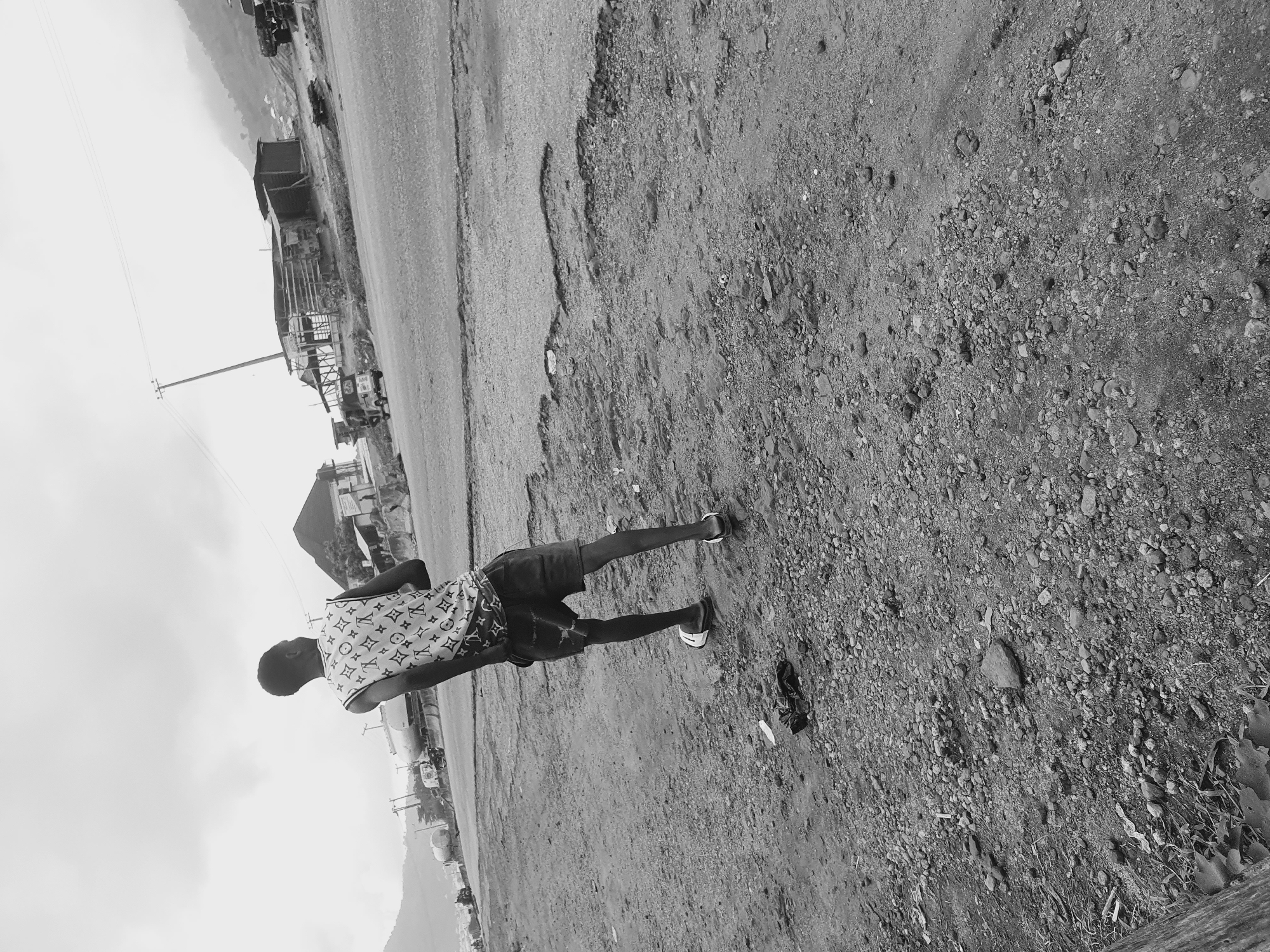 A black and white photograph shows a person standing on a dirt and gravel surface adjacent to a paved road. The person is wearing shorts and a short-sleeved shirt with a repeating pattern of "LV" logos. In the background, there are structures including buildings and utility poles. The background is blurry, suggesting some distance. The photograph appears to be taken at a slight angle. The location is Lokoja, Nigeria.