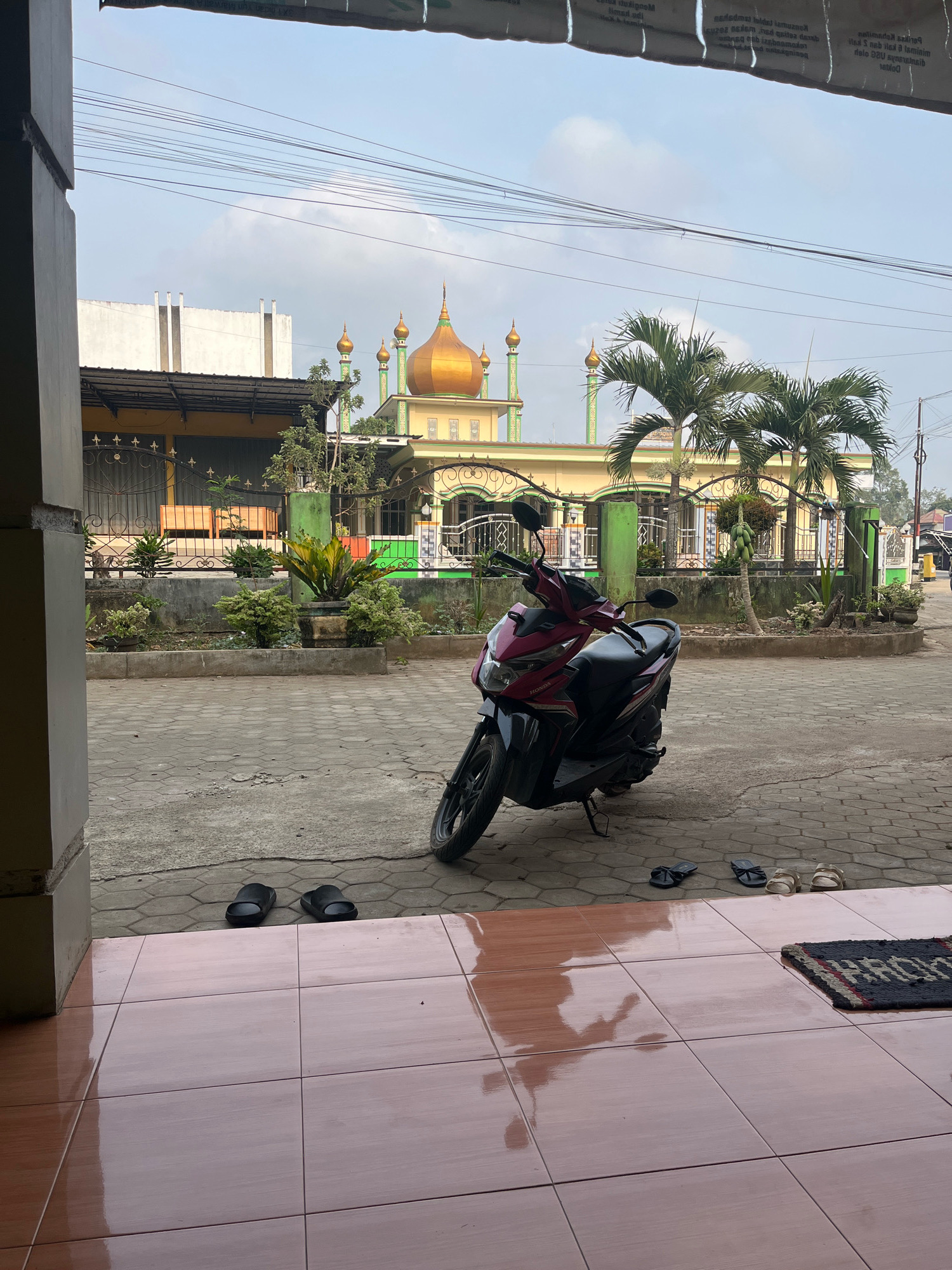 In Kemuning, Indonesia, a red and black motorcycle is parked on a patterned concrete driveway in front of a mosque. The mosque features a golden dome and several minarets, with palm trees and other greenery surrounding it. Two pairs of black flip-flops and two pairs of tan sandals are visible on the driveway. A small woven mat is placed on a tiled patio in the foreground. The sky is overcast, and the scene appears to be illuminated by daylight.