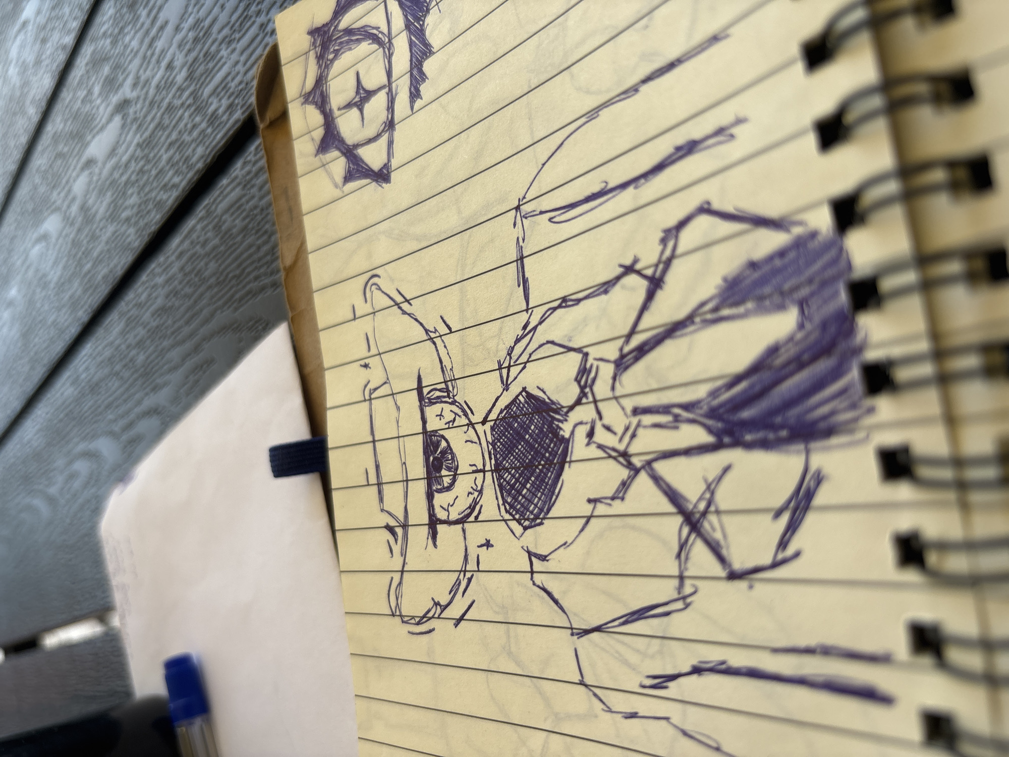 This is a close-up, angled, eye-level shot of a sketch in progress on a yellow, lined notepad. The sketch, drawn with a purple pen, appears to be of a character's face, possibly from an anime or manga. The subject is depicted in profile, with a large eye that is heavily cross-hatched, suggesting it is the focal point of the drawing. Other features like hair and part of a mouth are visible, also rendered with loose, energetic lines. The sketch is partially obscured by the spiral binding of the notepad and a portion of a blank white page, which is held in place by a dark blue elastic band. To the left of the notepad, a blurred view of a wooden surface, possibly a table or bench, is visible. A blue pen cap is also faintly visible at the bottom left of the frame. The scene is lit by natural light, suggesting it is daytime. The overall impression is of a casual, creative moment captured outdoors, perhaps in a park or on a porch, given the texture of the wooden surface. There is no discernible text in the image. The location context is Karviná, Czechia.
