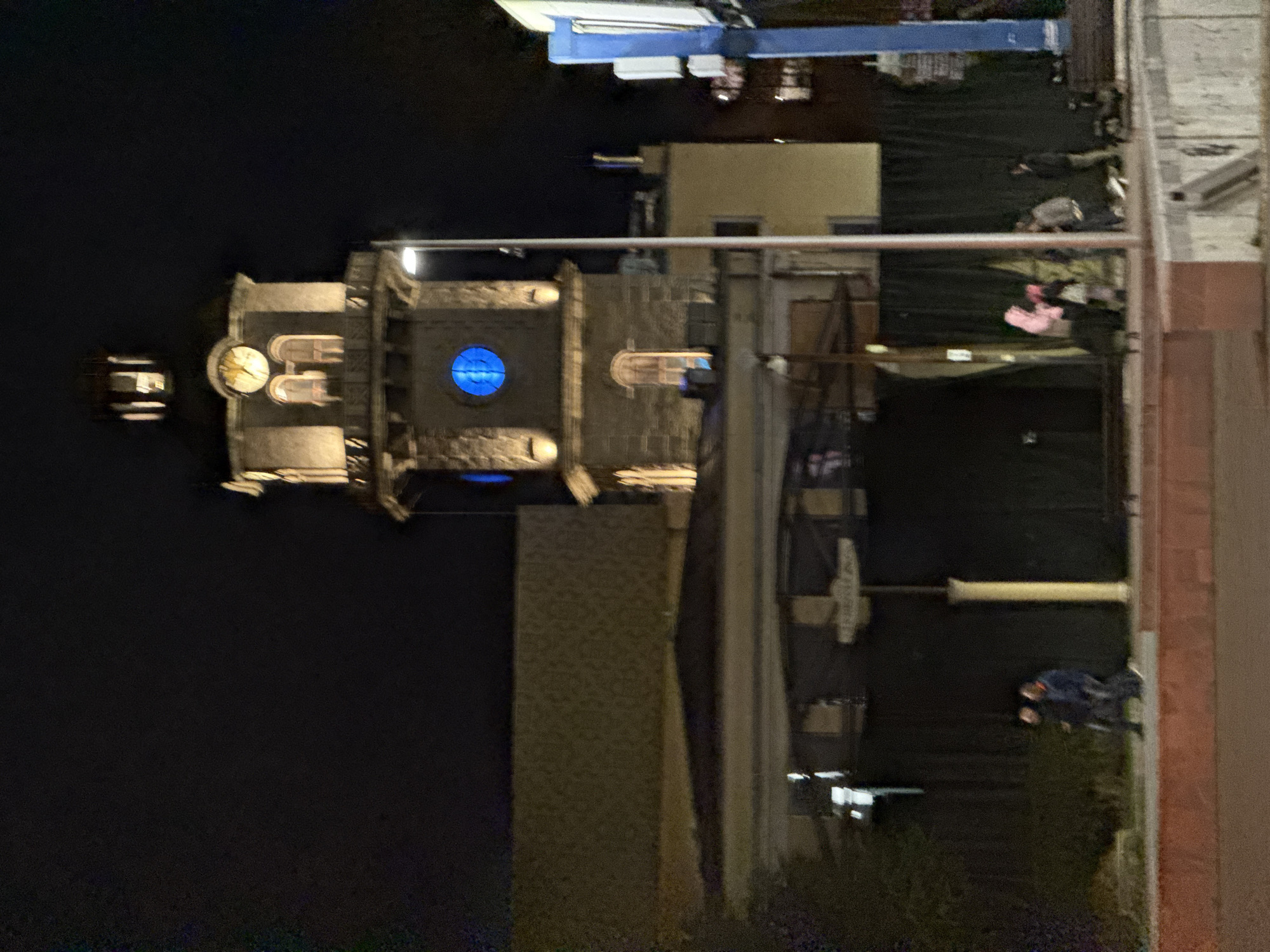 The image captures a nighttime urban scene centered on the Clock Tower (Часовникова кула) in Varna, Bulgaria, located on Knyaz Boris I Boulevard.
The dominant structure is a stone-built clock tower, illuminated with warm yellow light. Its upper section features a circular clock face emitting a yellow glow. Below this, two arched windows are visible, also lit with warm light. A prominent circular window located in the tower's mid-section glows bright blue. A third arched window is visible below the blue circular window.
To the right of the tower, a street light pole with a visible luminaire stands vertically. Further to the right, a segment of a blue and white vertical sign structure is present. Below the tower, a dark building facade, possibly covered or undergoing renovation, spans horizontally; a single cream-colored column supports this structure.
In the foreground, a raised reddish-brown pavement or walkway is visible. On the left side of this pavement, two individuals are walking from left to right, obscured by blur. One wears dark clothing; the other has a dark jacket with a reddish accent. On the right side of the pavement, a group of approximately five individuals is gathered. One person in this group is wearing a distinct bright pink hoodie. The clothing of the other individuals in the group varies from dark to light casual wear. The nature of their interaction is unclear due to blur. A low concrete barrier is positioned near this group. To the far right, a partial view of a lone individual is visible. The sky above is dark.