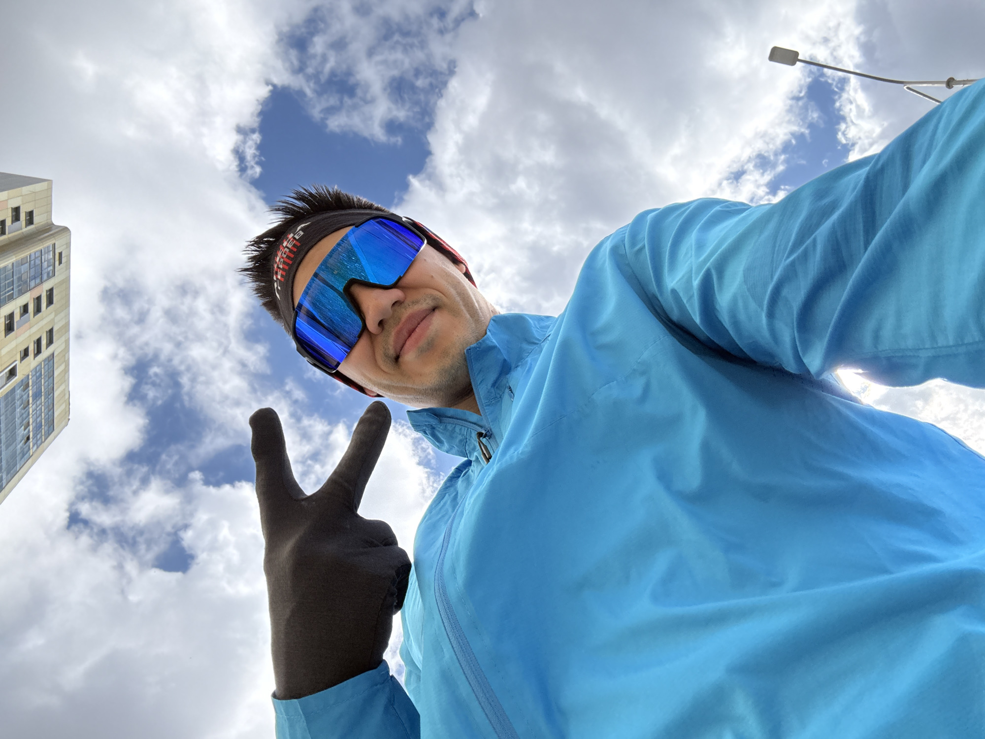 A low-angle selfie captures a person looking towards the camera, making a peace sign with their gloved left hand. The individual wears a bright light blue jacket, black athletic sunglasses with reflective blue lenses, and a black headband featuring white and red text, which appears to be "SCHEELS." His short, dark hair is visible above the headband, and a subtle smile plays on his lips. The background is dominated by a cloudy sky, showcasing a mix of white and grey clouds with distinct patches of blue sky peeking through.
On the far left, a tall, light-colored modern building with numerous windows can be seen, suggesting an urban environment. To the far right, a slender street light pole with a small rectangular light fixture extends into the sky. The scene takes place during the daytime under partially cloudy weather conditions. This image provides a glimpse of an urban setting in Kazan, Russia.