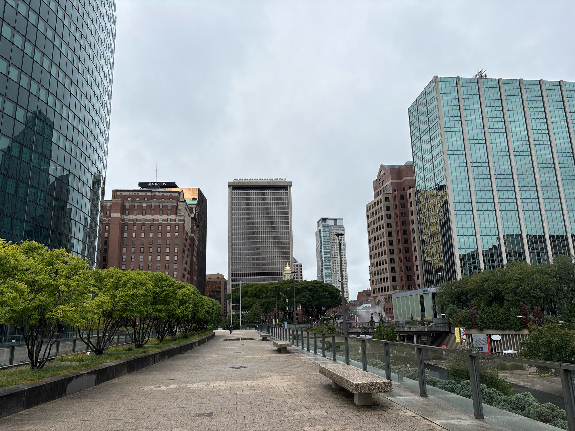 In Hartford, Connecticut, United States of America, an overcast day reveals a city park walkway with benches and trees. A wide, paved walkway, constructed with tan brick pavers, stretches into the distance, bordered by a low, dark planter box filled with green grass and trees. To the left, a tall, modern building with a curved facade of blue-tinted glass windows reflects the sky. Across the walkway and to the right, several other buildings rise, including a red brick building with numerous small windows, a grey, rectangular skyscraper with many windows, and a taller, blue-tinted glass skyscraper with vertical white trim. A clear glass railing with silver posts separates the walkway from a lower level of the park, which features more greenery and some exposed concrete structures. Two concrete benches are situated on the walkway, facing away from the viewer. A single person is visible in the distance, walking away from the camera. The overall atmosphere is one of an urban, pedestrian-friendly environment on a cloudy day.