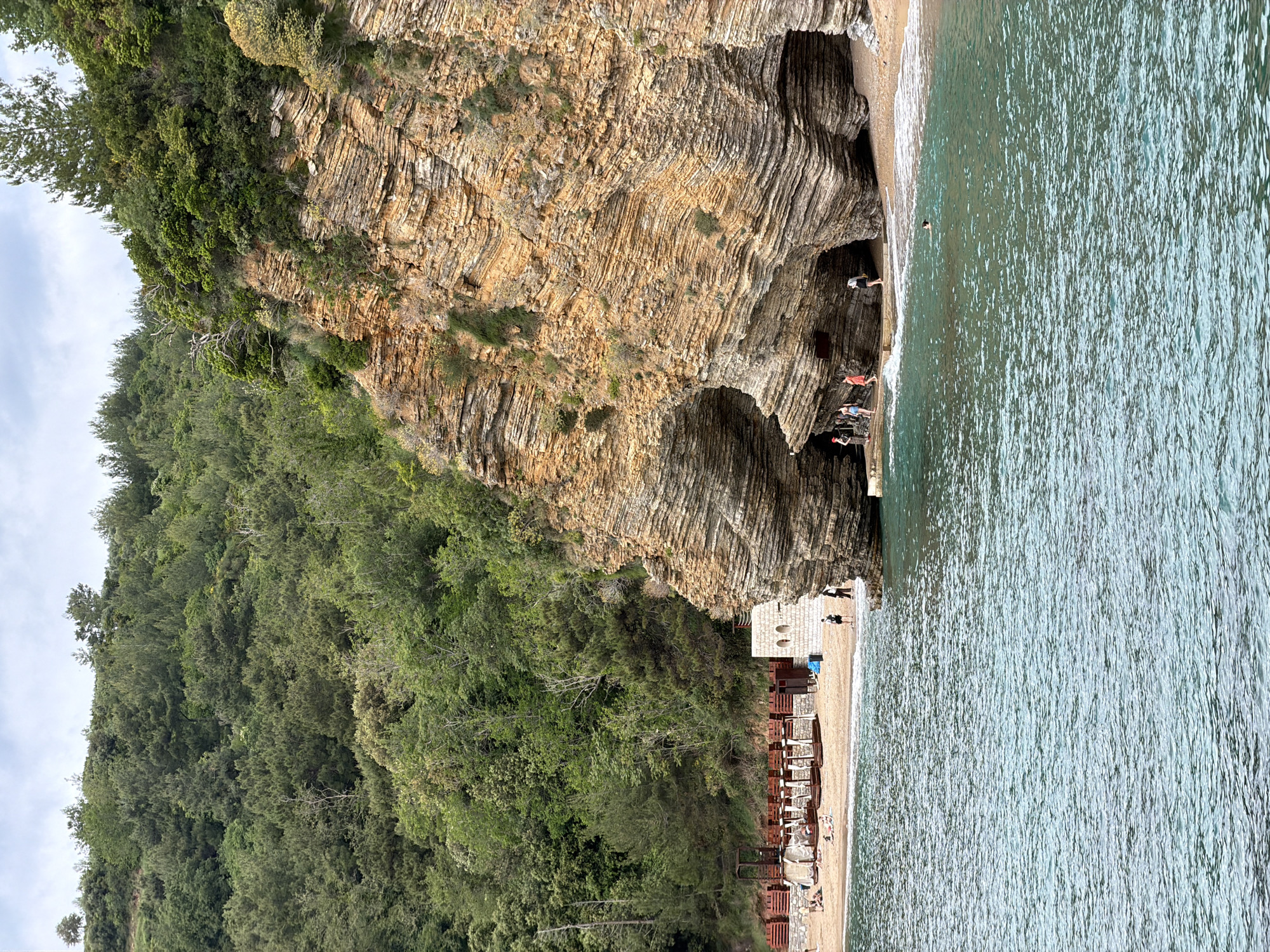 The image depicts a coastline scene in Budva, Montenegro. A large, layered rock formation dominates the view, with several caves and crevices visible at its base. A sandy beach stretches along the coastline, equipped with numerous lounge chairs and umbrellas. Several people are present, some standing near the caves and others relaxing on the beach. The sea occupies a significant portion of the image, with its surface reflecting the sunlight. Dense green vegetation covers the top and left of the rock formation and extends up the cliffside.