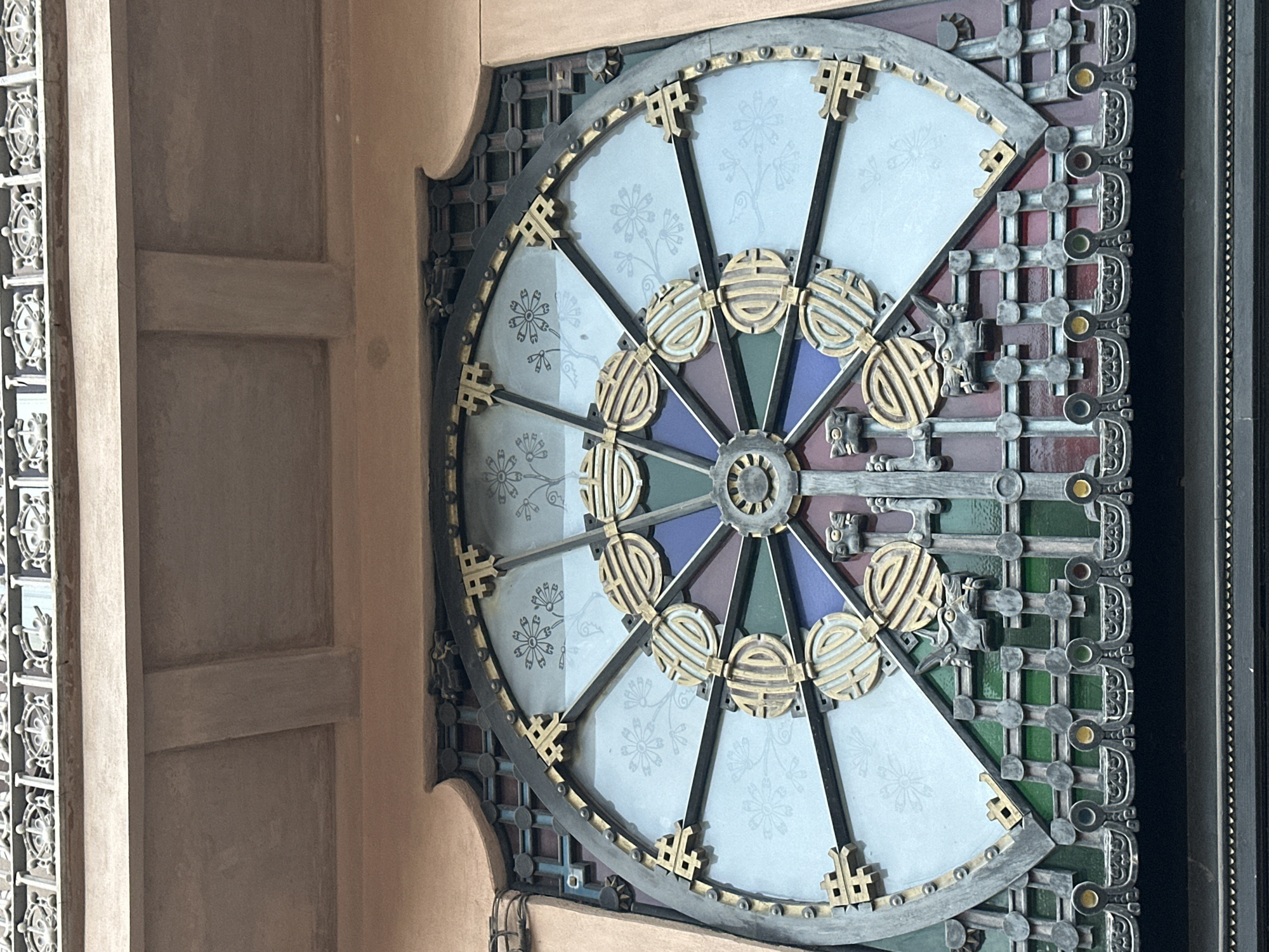 This is a medium shot, eye-level, vertical, exterior photograph of an ornate circular window. The window is framed by a dark metal structure that appears to be part of the building's facade. The window itself is a large stained-glass design featuring a central hub with radiating spokes. The spokes are adorned with golden, intricate, circular motifs that resemble Chinese characters or symbols, and small, stylized floral patterns are etched into the translucent glass panes. The lower portion of the window incorporates colorful stained glass in shades of green, blue, and red. Below the circular window, there is a decorative metal grille with a repeating pattern of circular elements and what appear to be stylized dragon heads. The building facade around the window is a muted pinkish-tan color, and above the window, a portion of an upper architectural element is visible, including a projecting ledge and a decorative metal railing. The overall aesthetic suggests a building with an East Asian influence, possibly a temple or a building designed to evoke such a style. The lighting is even, indicating it is daytime with diffused light. There are no people or animals visible in the image. This architectural element is located at Casa Batlló in Barcelona, Spain.