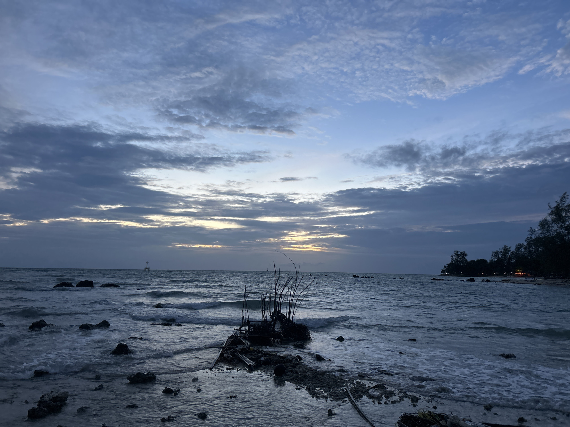 The image depicts a tranquil twilight scene at a beach in the Ko Pha-ngan District, Thailand. The sky is filled with a blend of blues and grays, with wispy clouds and a subtle hint of golden light near the horizon, suggesting either sunrise or sunset. The sea is calm, with gentle waves lapping at the shore, creating a frothy white edge. Scattered rocks dot the waterline and are submerged in the water. In the foreground, a clump of dark, tangled debris, possibly driftwood and vegetation, rests on the wet sand, with a few thin branches reaching upwards. In the distance, a small, white lighthouse-like structure stands on the water. The far shoreline is lined with dark trees, and a few distant lights indicate human presence. The overall atmosphere is serene and natural, with the fading light casting a subdued and somewhat melancholic mood. There are no people visible in the image.